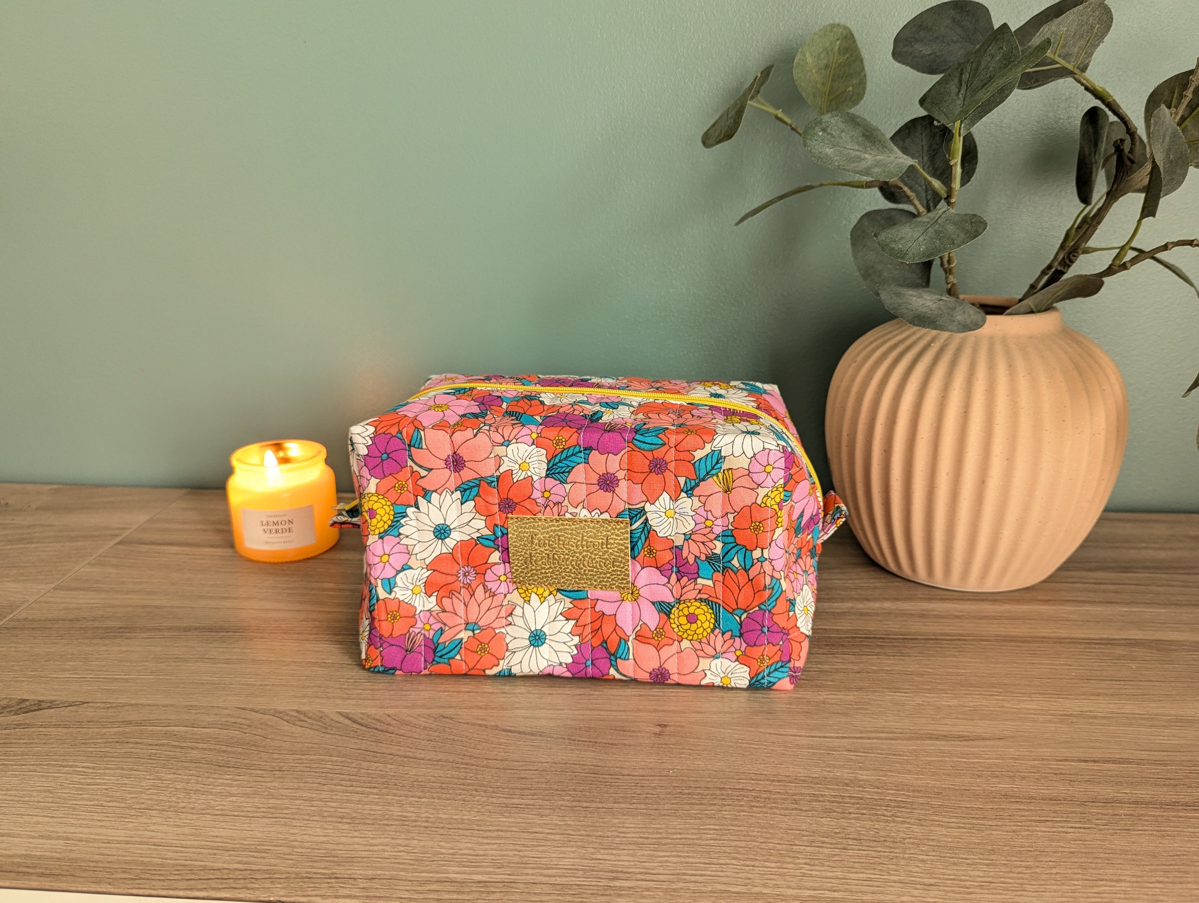A colorful floral makeup bag sits on a wooden surface, accompanied by a small candle and a potted plant.