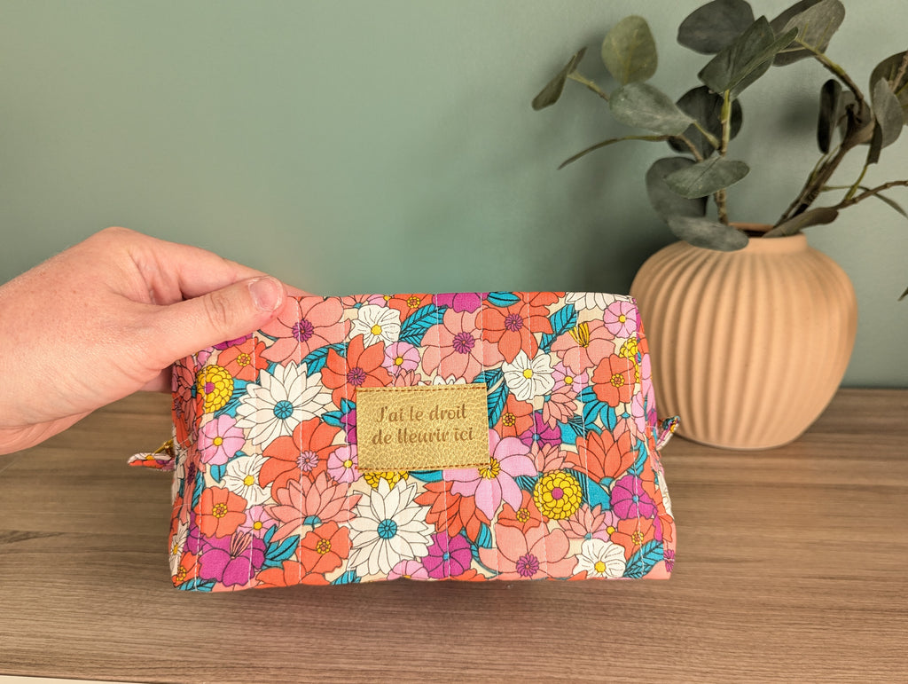 A hand holding a colorful floral makeup bag with a gold-colored tag that says "Jat le droit de la Heure vie".