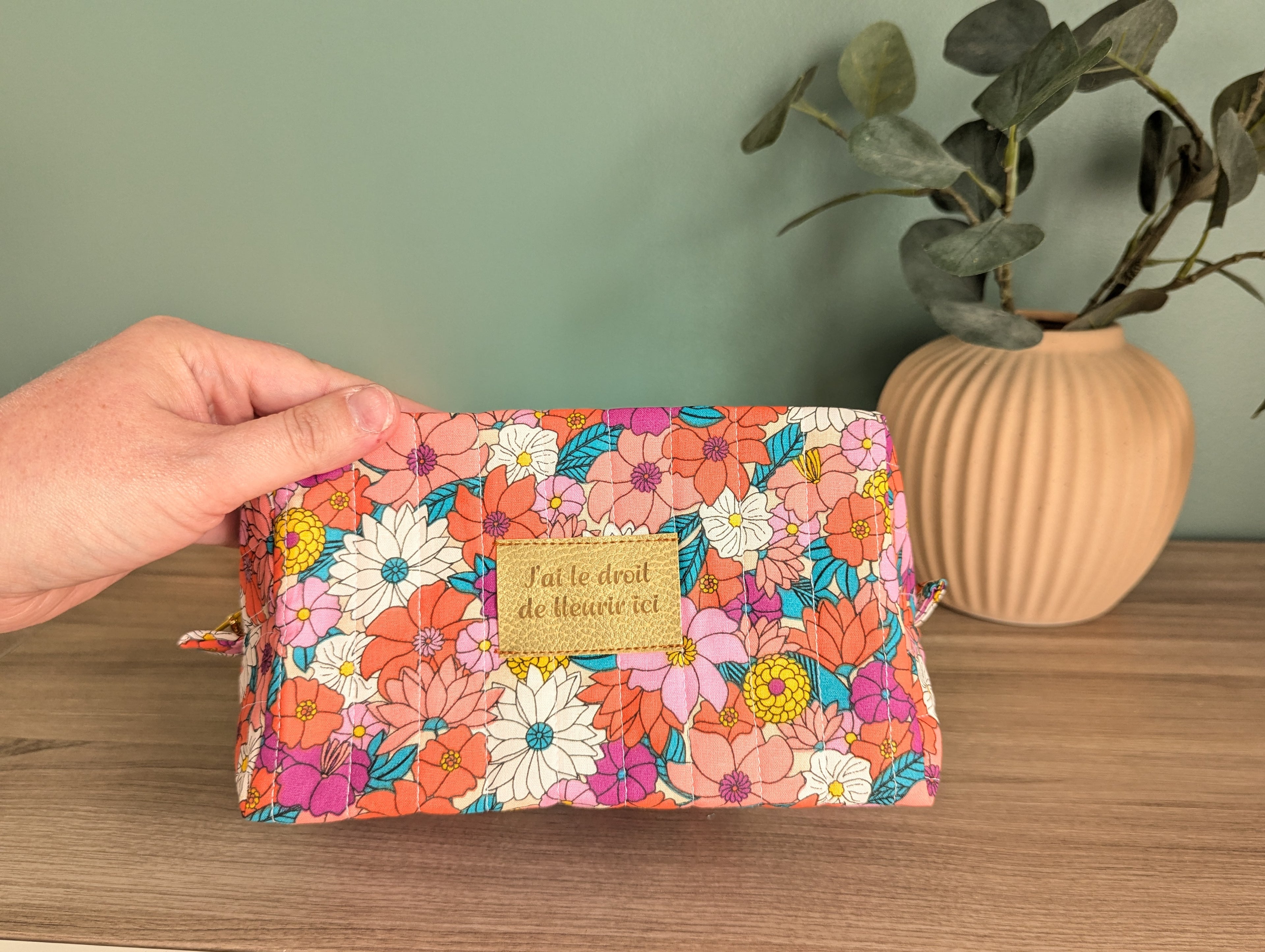 A hand holding a colorful floral makeup bag with a gold-colored tag that says "Jat le droit de la Heure vie".