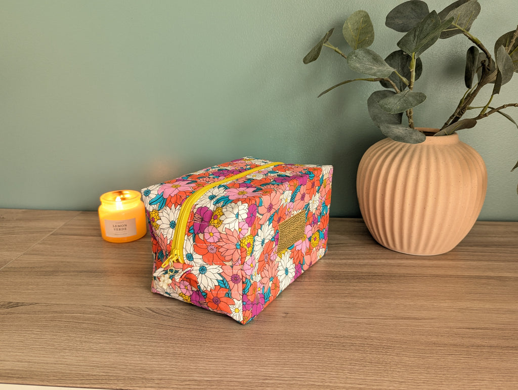 A floral-patterned cosmetic bag sits on a wooden surface next to a candle and a potted plant, creating a cozy and inviting atmosphere.