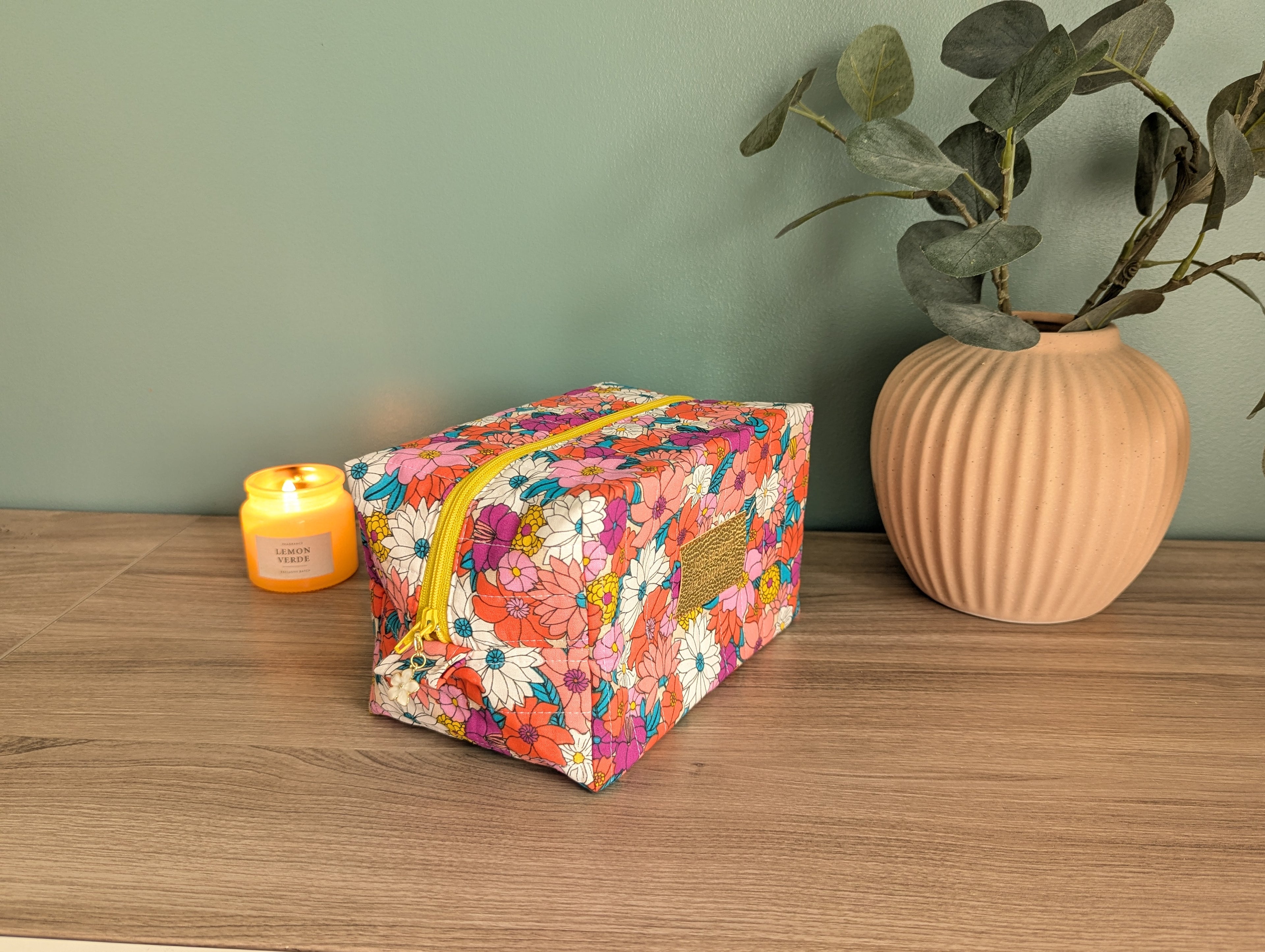 A floral-patterned cosmetic bag sits on a wooden surface next to a candle and a potted plant, creating a cozy and inviting atmosphere.