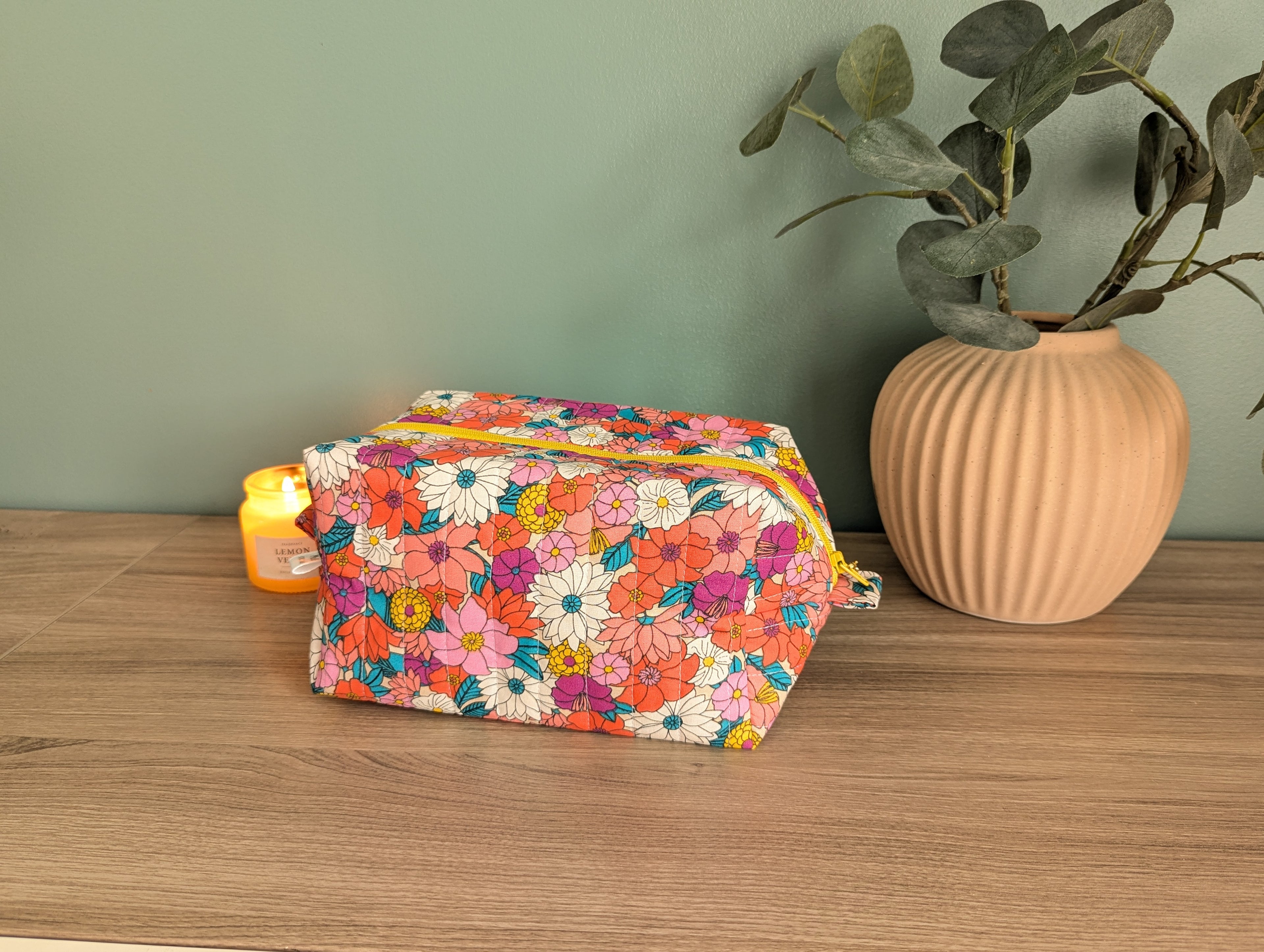 A colorful floral bag sits on a wooden surface next to a vase with green leaves, with a small yellow candle in the background.
