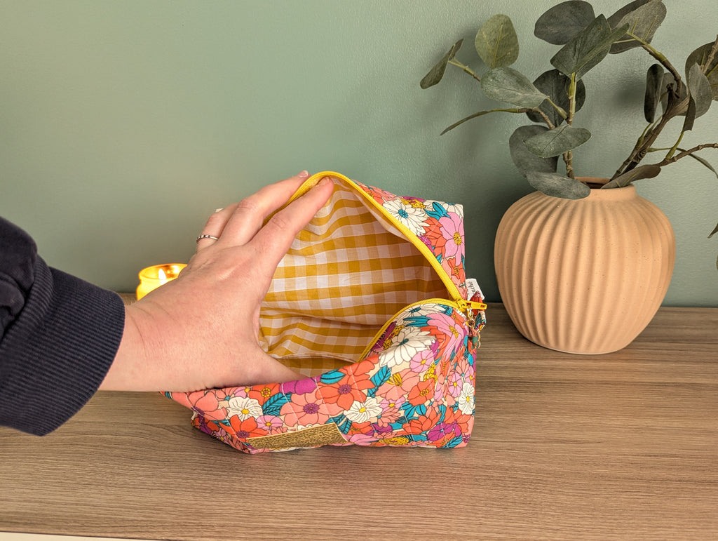 A hand is holding an open colorful pouch with a floral pattern, placed on a wooden surface next to a vase with green leaves.
