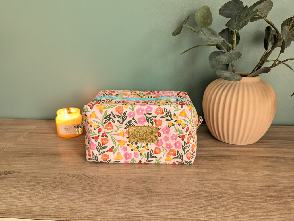 A floral-patterned cosmetic bag sits on a wooden surface, accompanied by a lit candle and a potted plant.