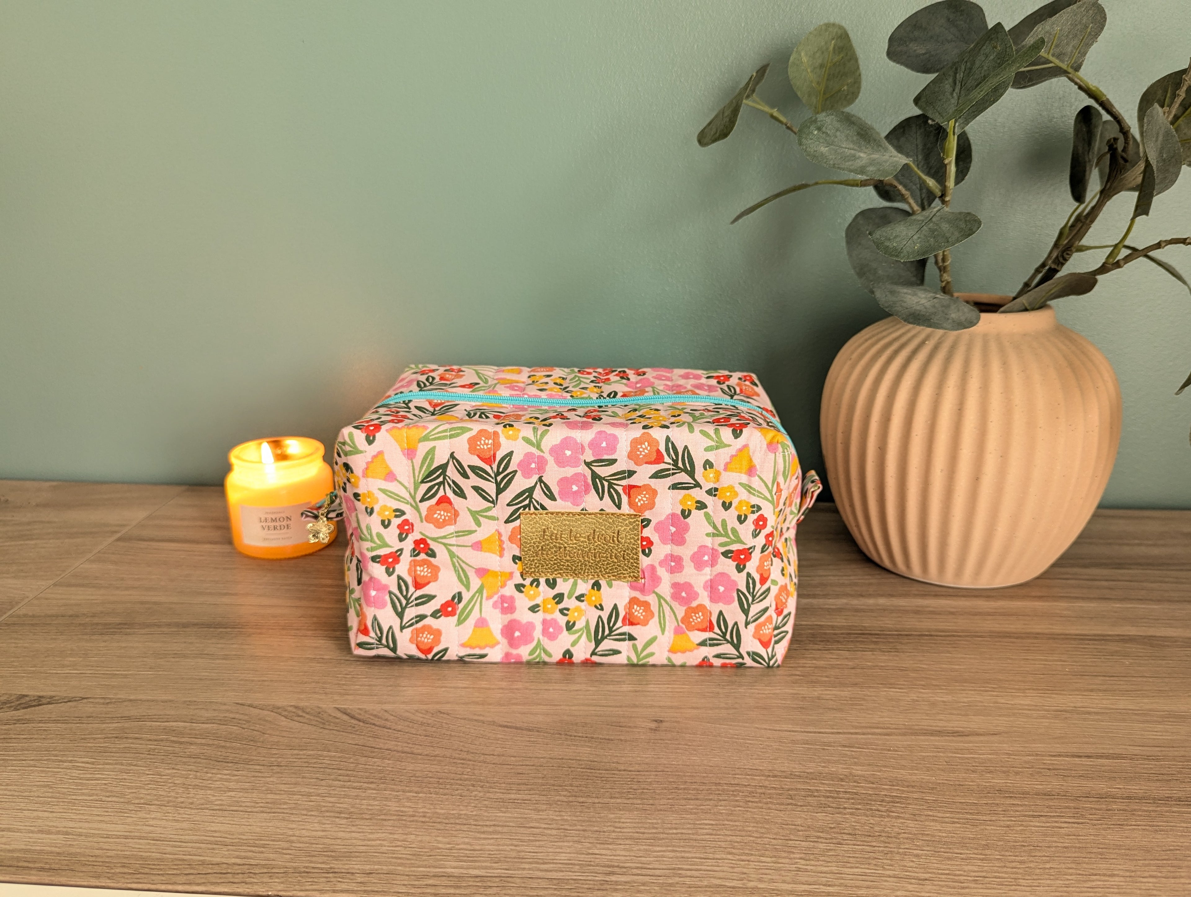 A floral-patterned cosmetic bag sits on a wooden surface, accompanied by a lit candle and a potted plant.