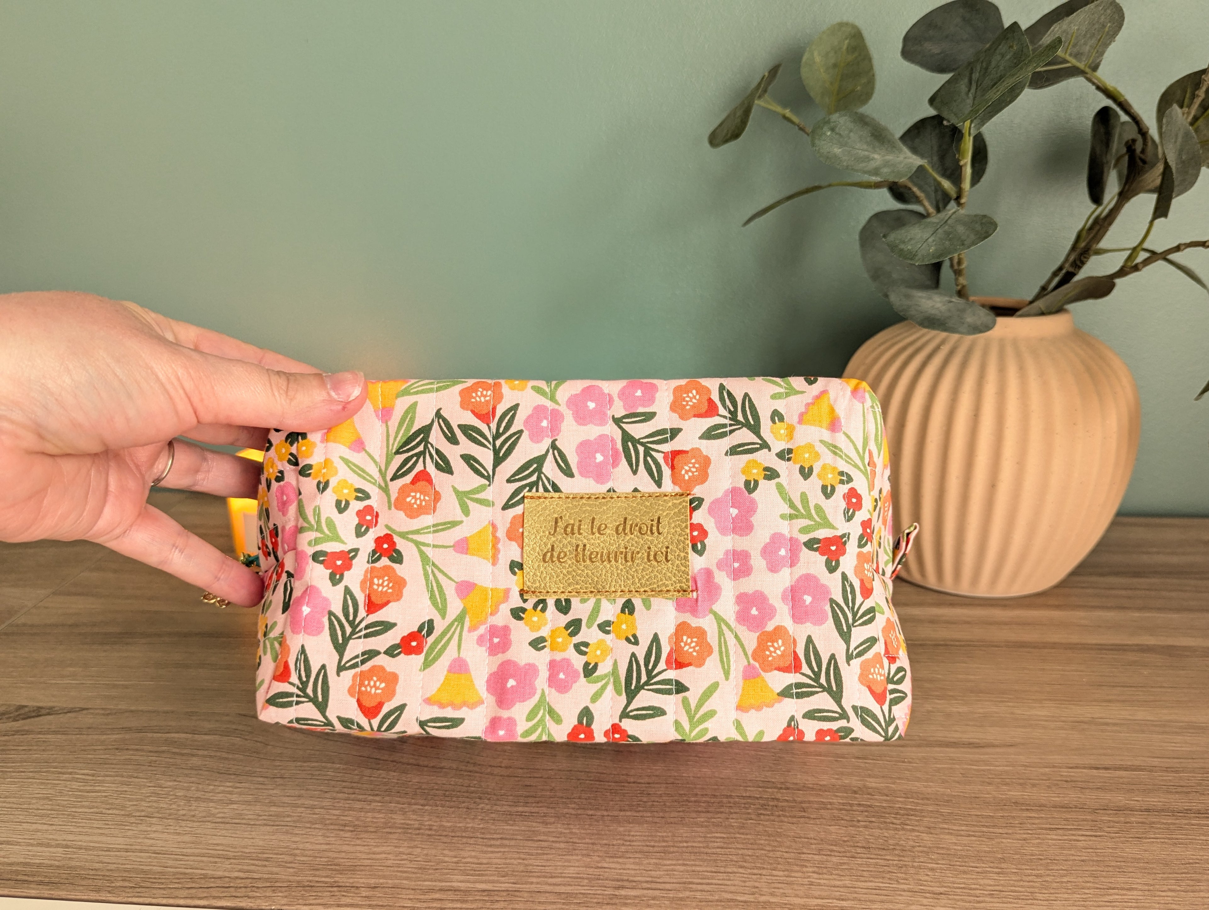 A hand holding a colorful floral makeup bag with a gold-colored label, placed on a wooden surface next to a potted plant.