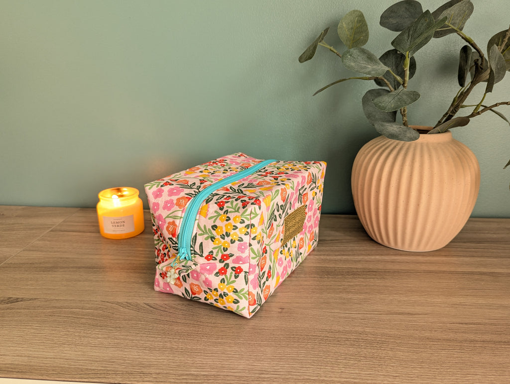 A floral-patterned cosmetic bag with a zipper sits on a wooden surface, accompanied by a small yellow candle and a potted plant.