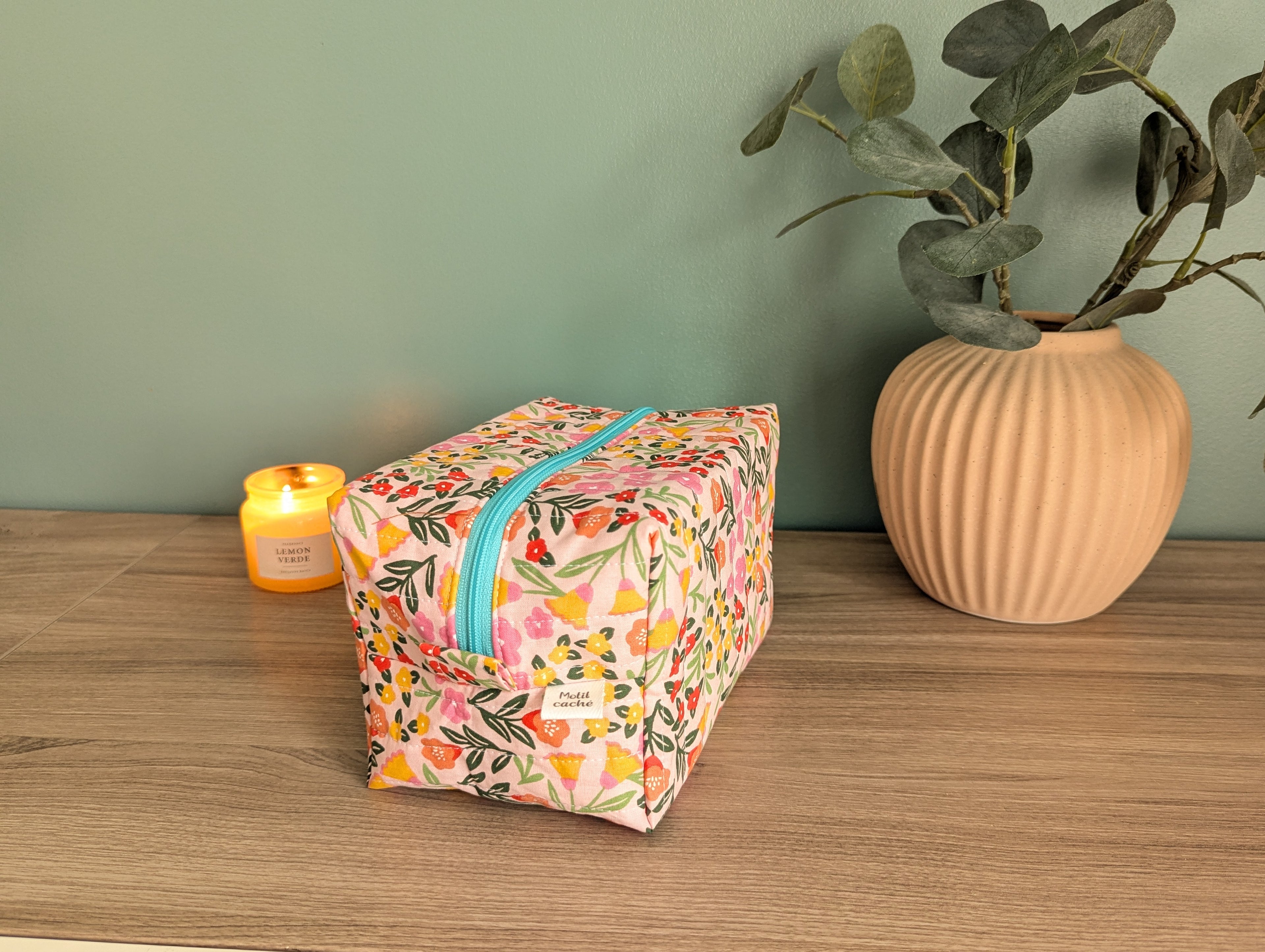 A floral-patterned makeup bag with a zipper sits on a wooden surface, accompanied by a small yellow candle and a potted plant.