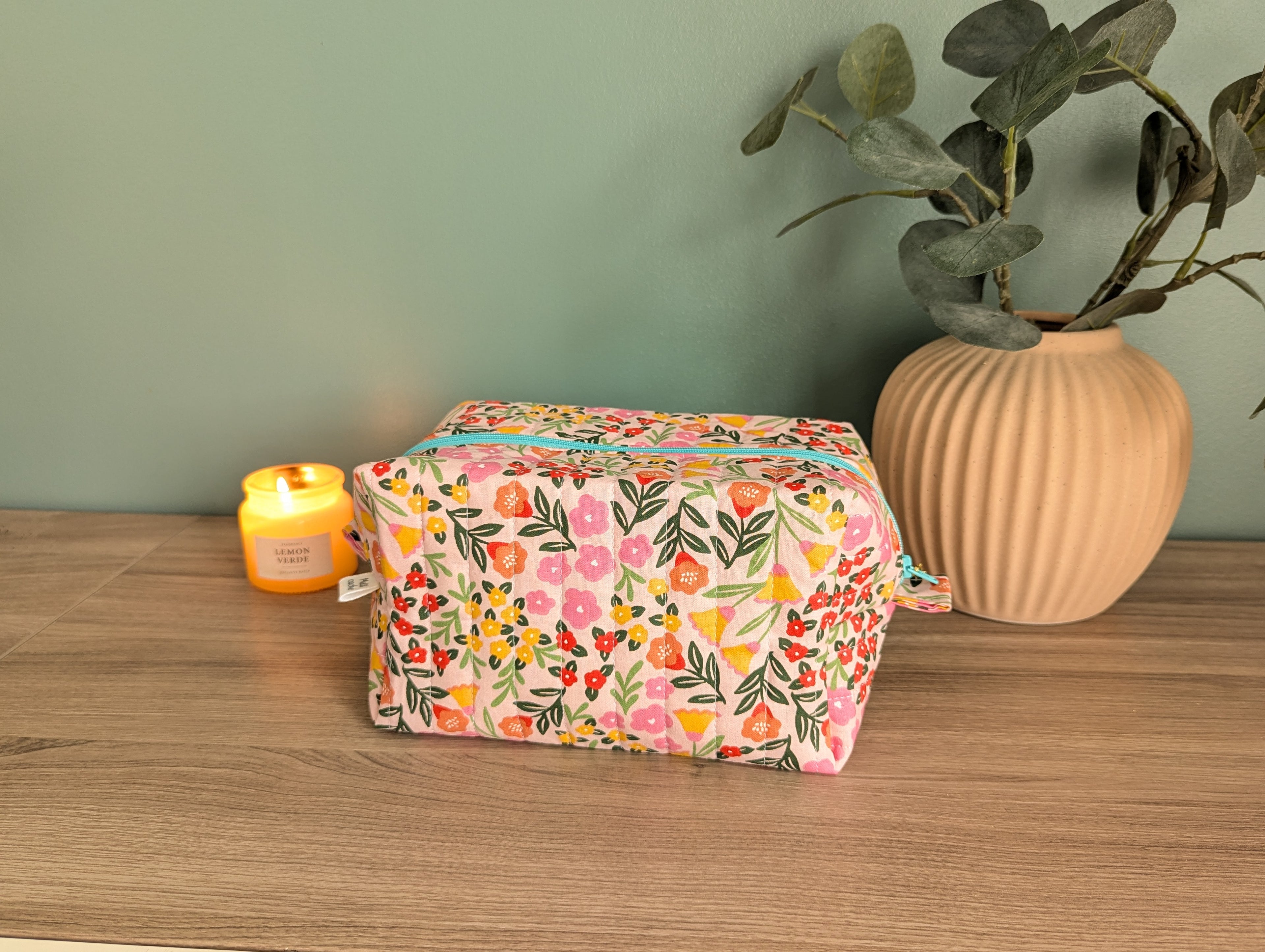 A floral patterned cosmetic bag sits on a wooden surface, accompanied by a lit candle and a potted plant.