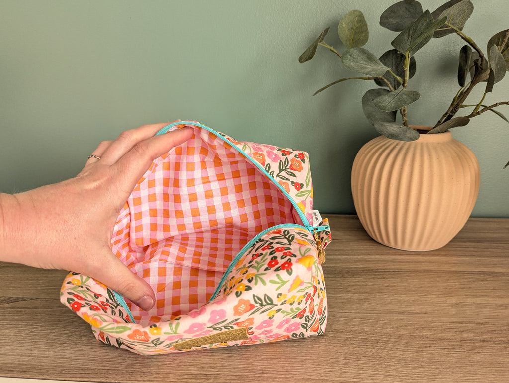 A hand is holding an open floral patterned bag on a wooden surface, with a potted plant and a vase in the background.