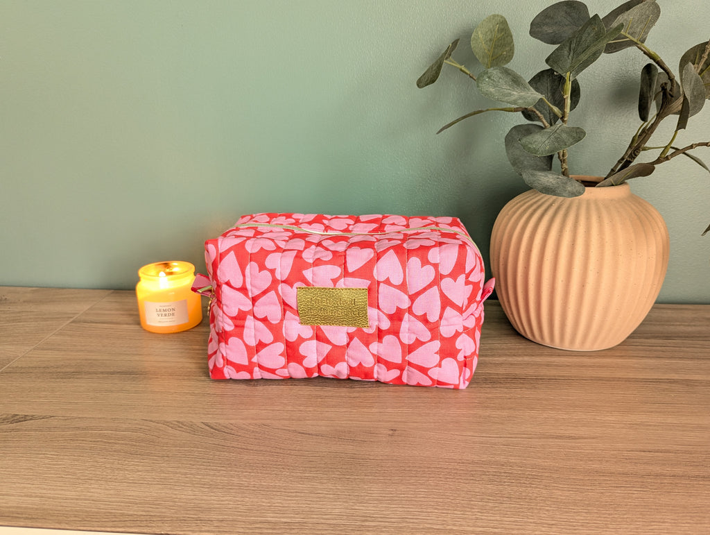A red and pink heart-patterned cosmetic bag sits on a wooden surface, accompanied by a small yellow candle and a potted plant.
