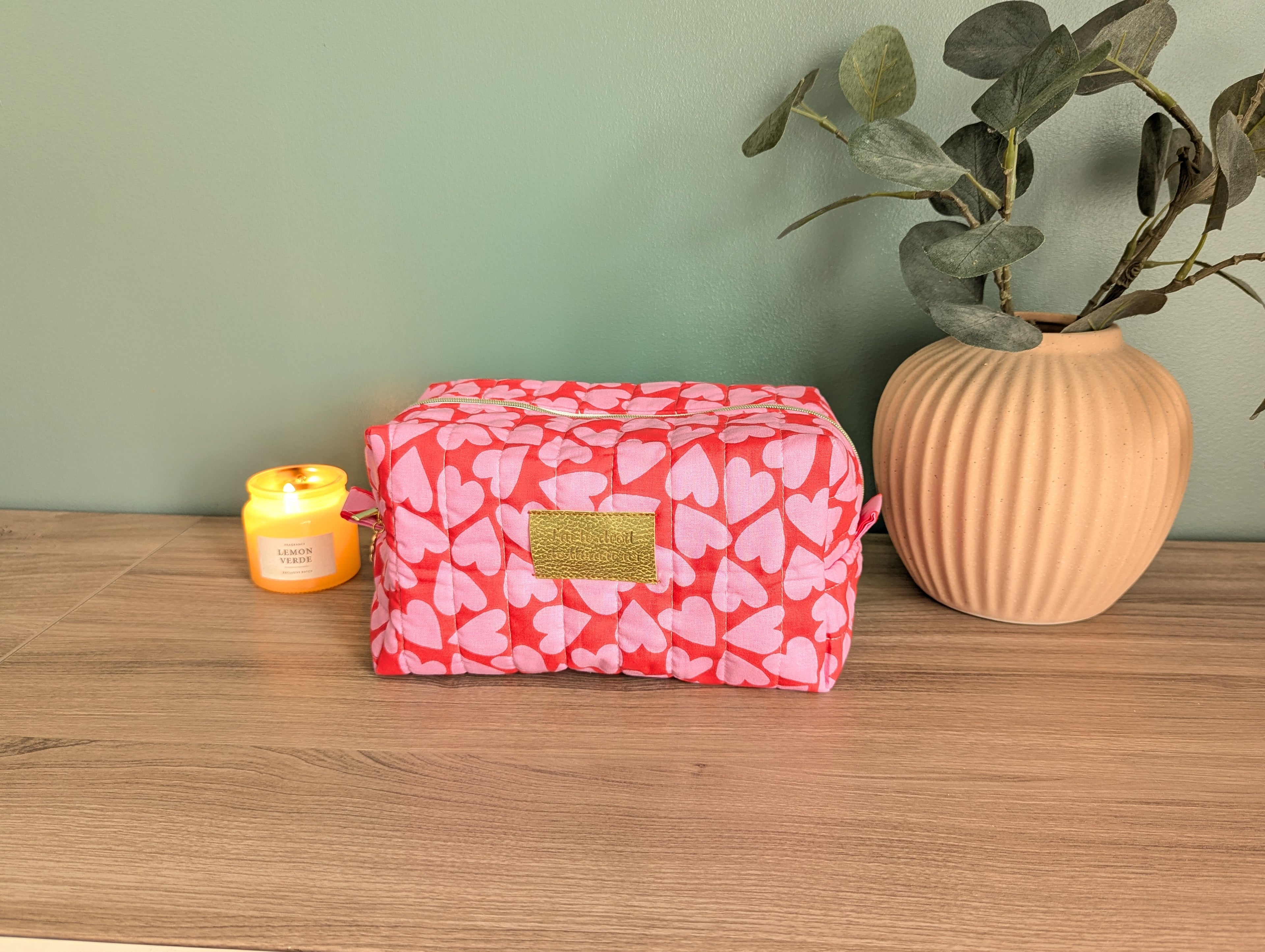 A red and pink heart-patterned cosmetic bag sits on a wooden surface, accompanied by a small yellow candle and a potted plant.