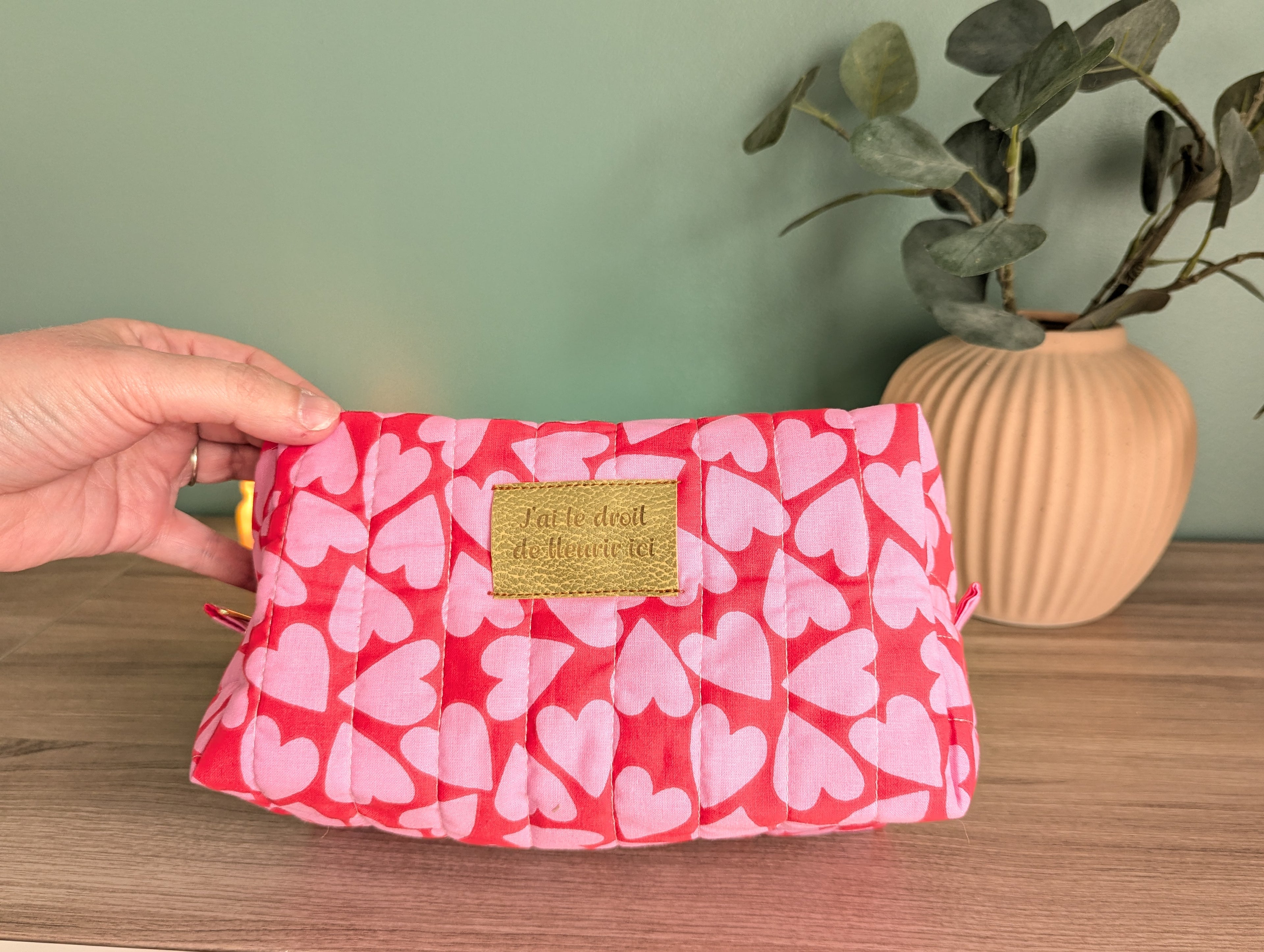 A hand is holding a red and white heart-patterned bag with a gold label that says "I love you". The bag is placed on a wooden surface, with a potted plant and a vase in the background.