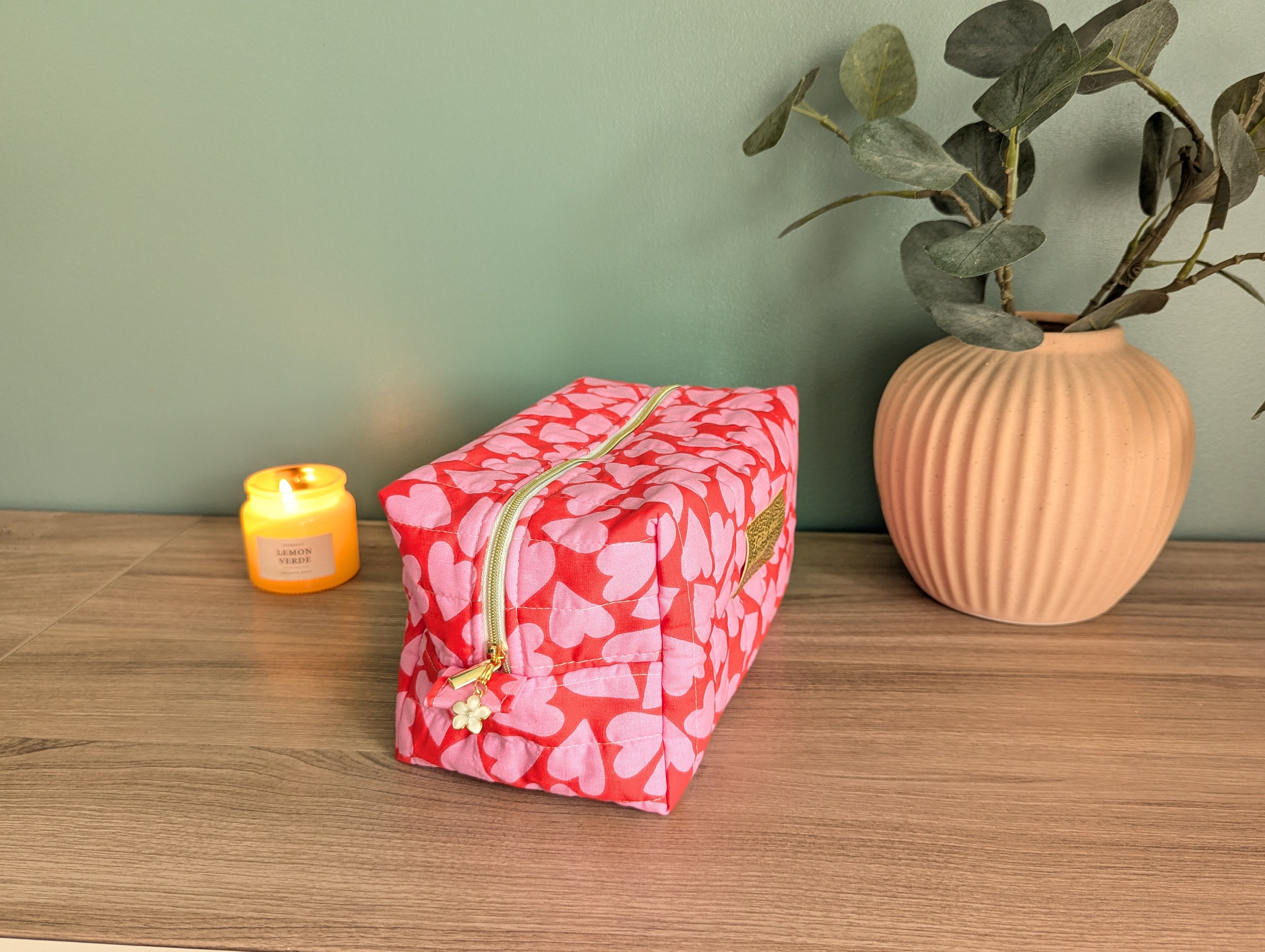 A red and pink heart-patterned cosmetic bag sits on a wooden surface next to a small yellow candle and a potted plant.
