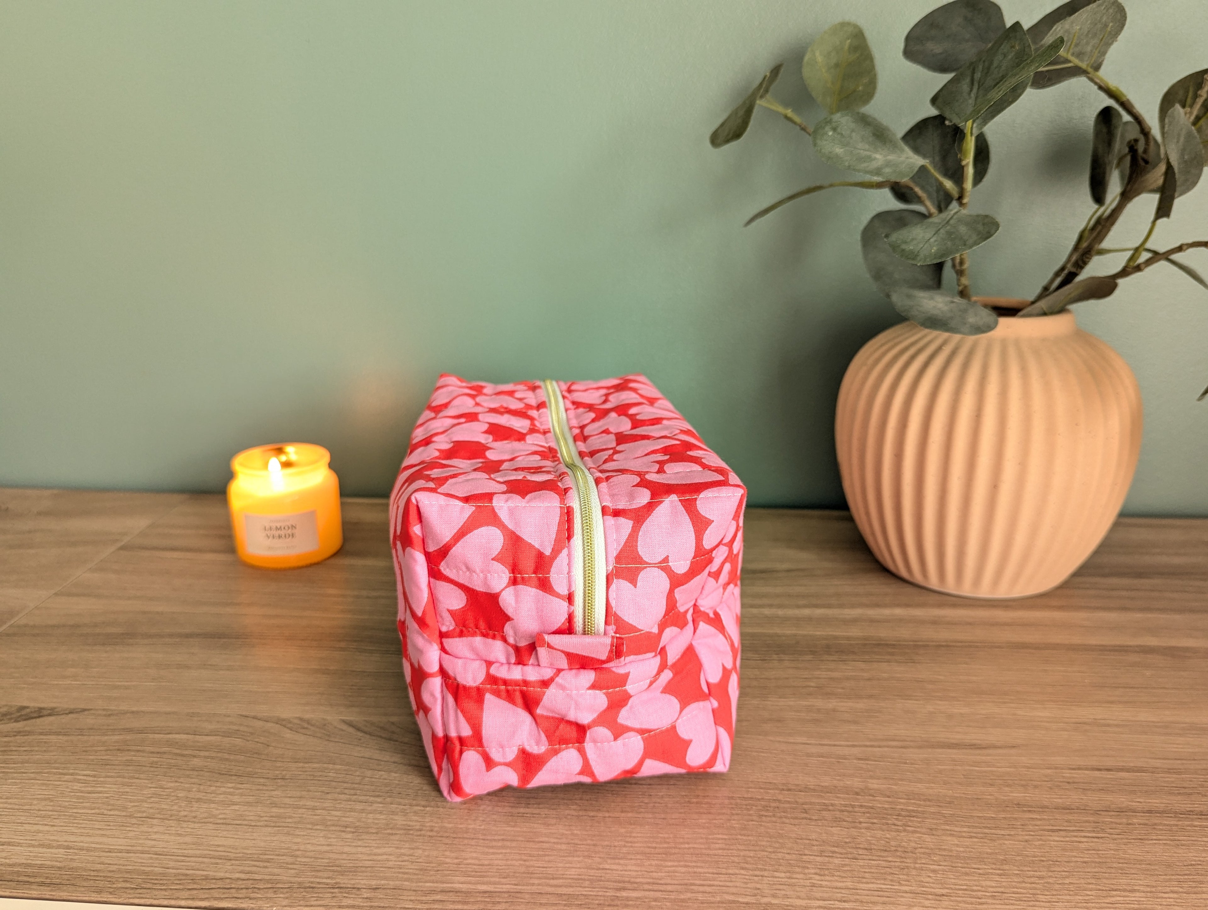 A pink and red heart-patterned bag sits on a wooden surface, accompanied by a small yellow candle and a potted plant.