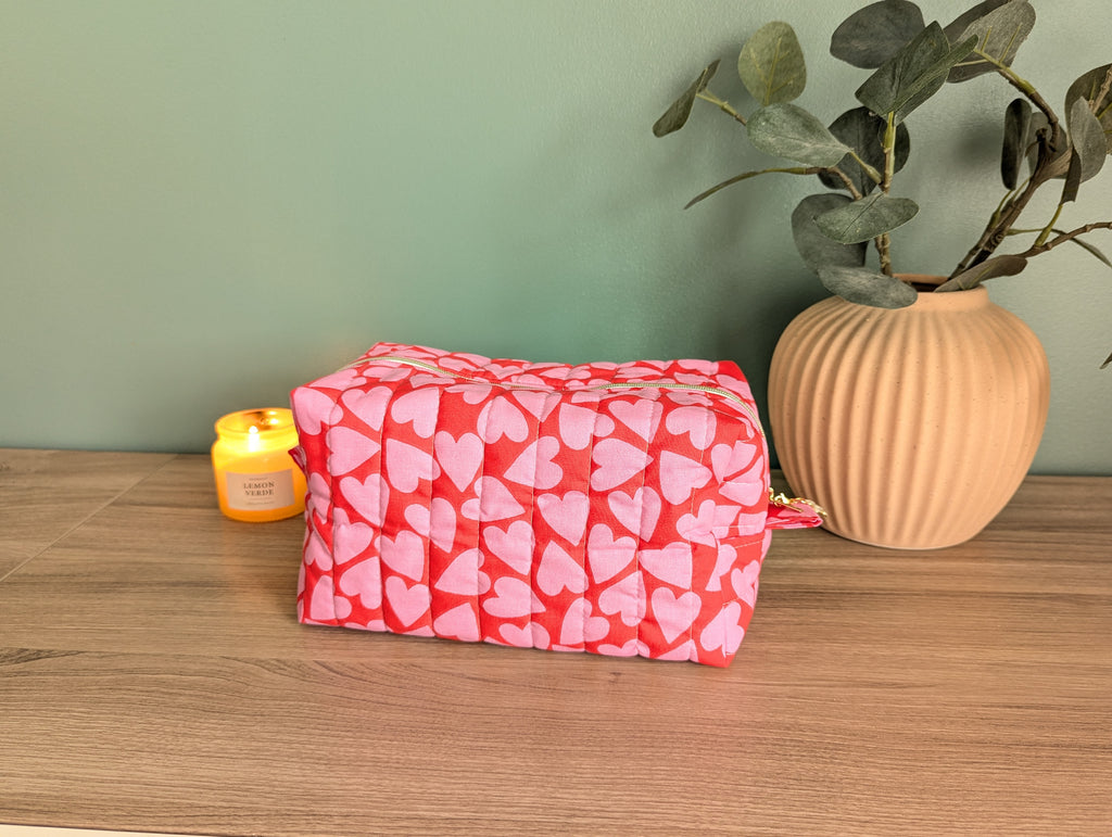 A red and pink heart-patterned bag sits on a wooden surface, accompanied by a small yellow candle and a potted plant.