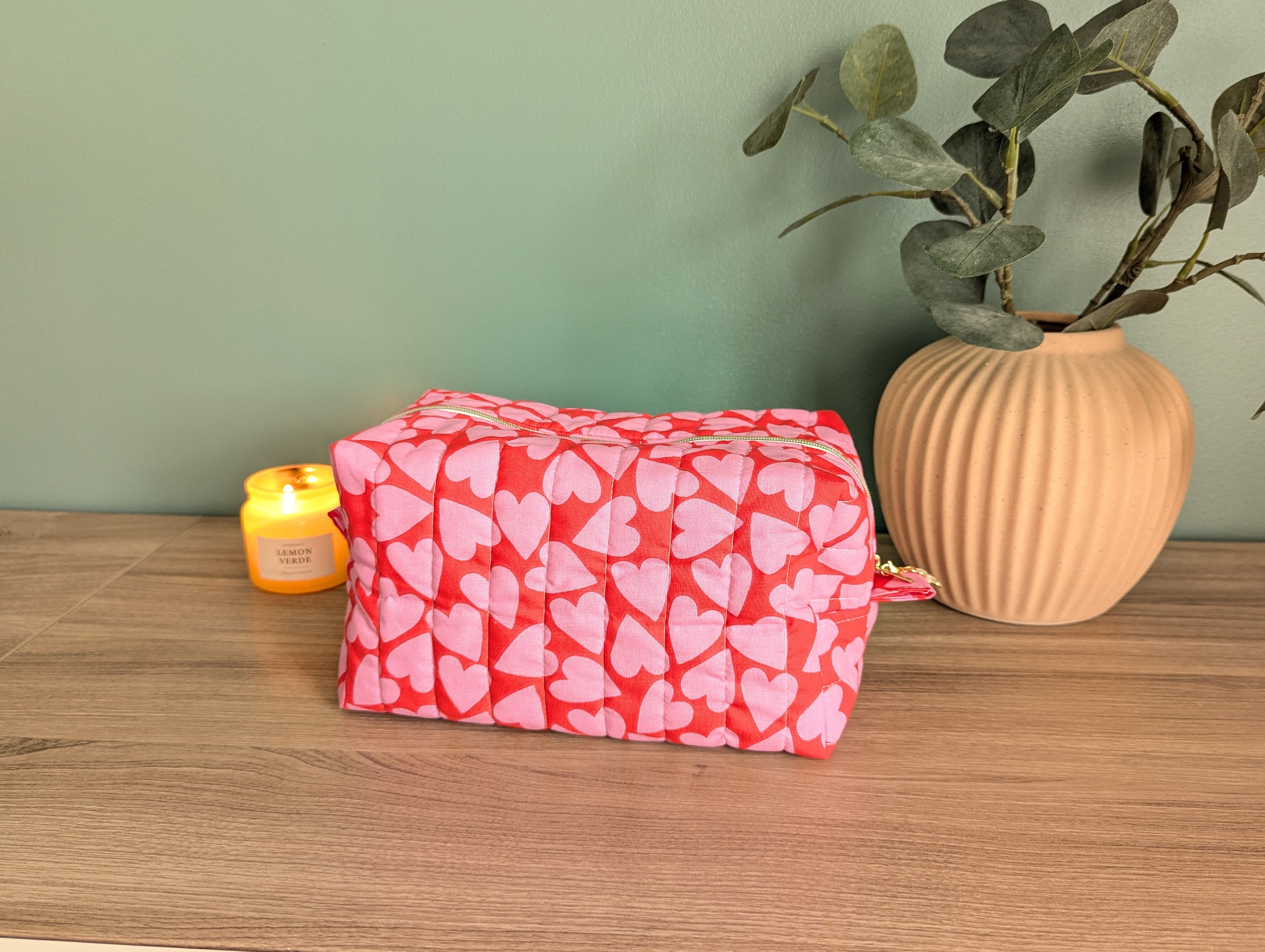 A red and pink heart-patterned bag sits on a wooden surface, accompanied by a small yellow candle and a potted plant.