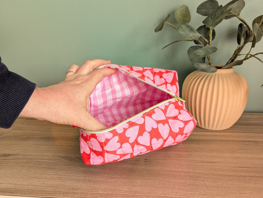 A hand is holding an open red and white striped cosmetic bag with a heart pattern on it, placed on a wooden surface next to a potted plant.