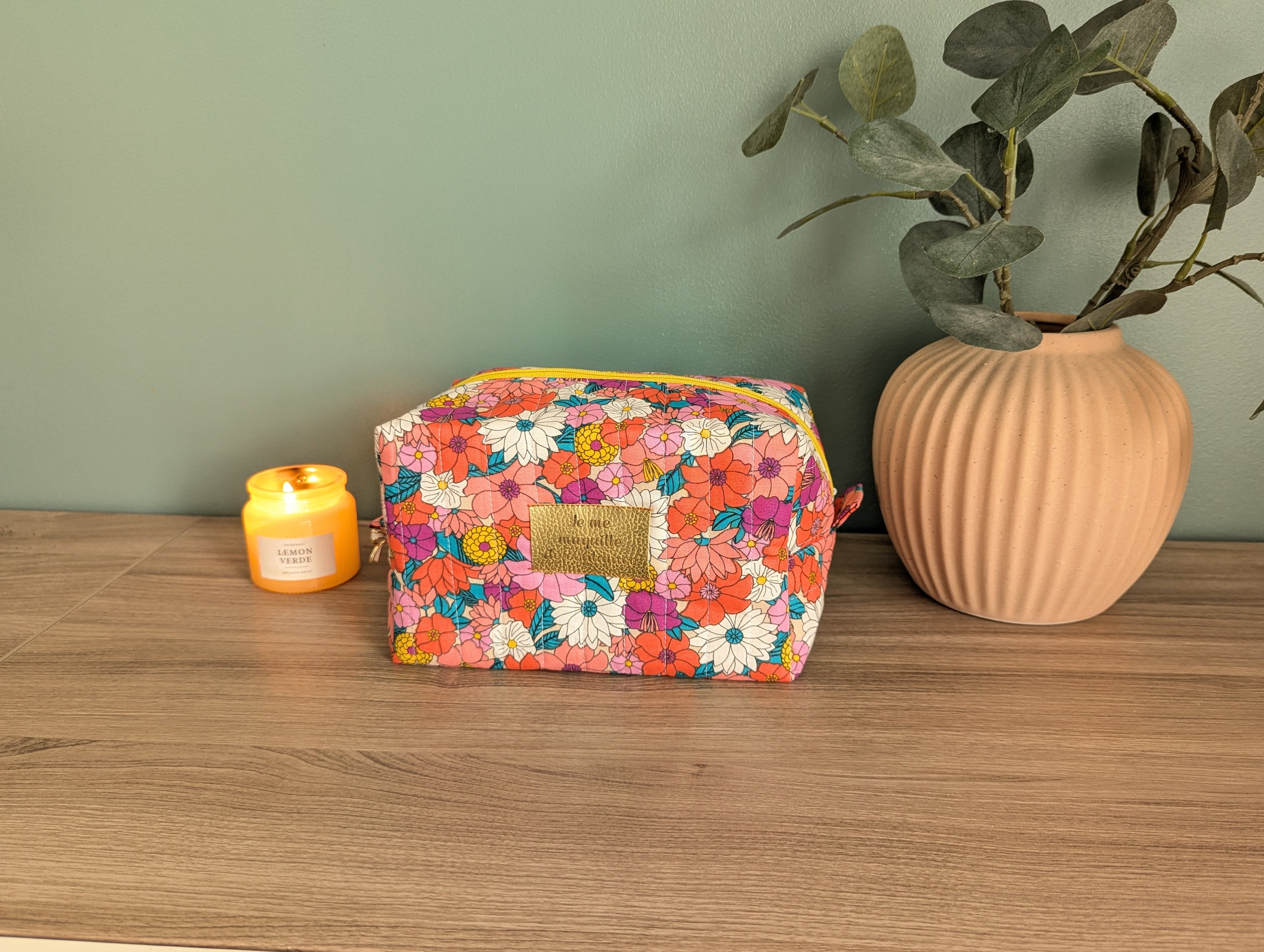 A floral-patterned makeup bag sits on a wooden surface next to a small yellow candle and a beige vase with green leaves.
