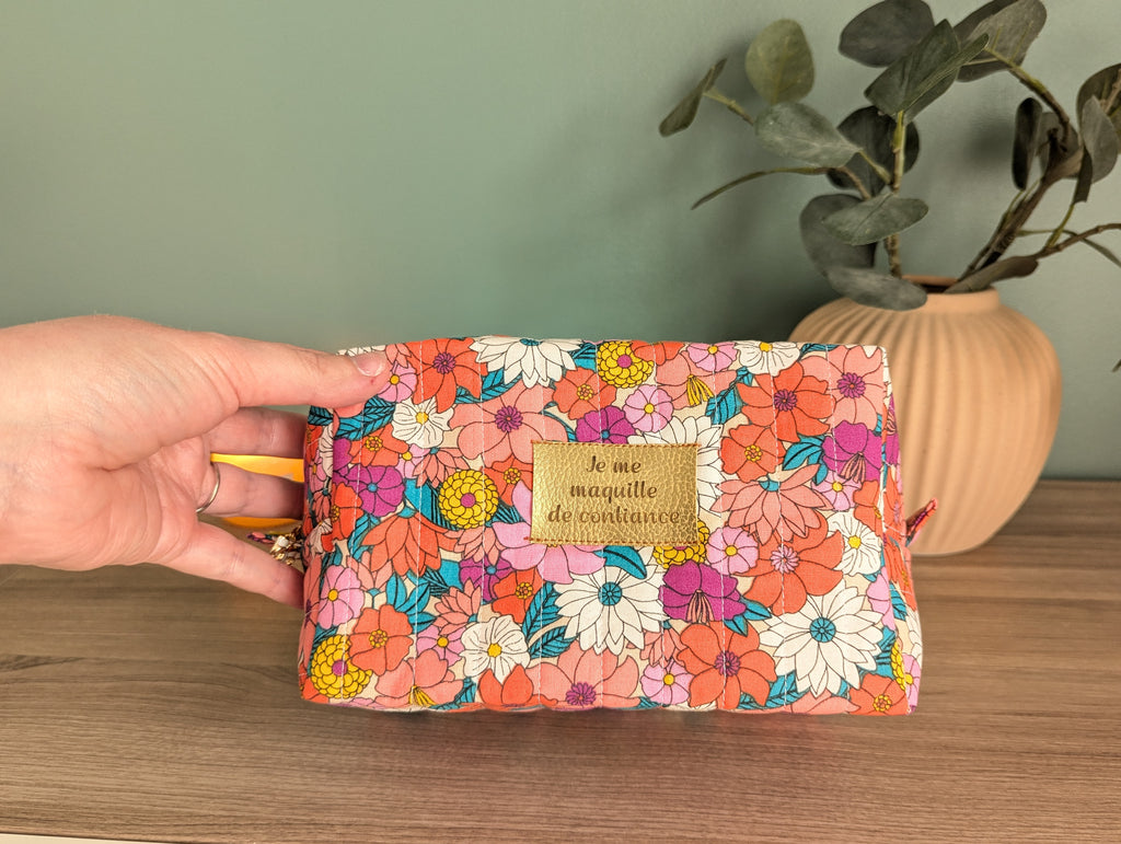 A hand holding a colorful floral makeup bag with a gold-colored tag that says "Je me mouquette de contenu".