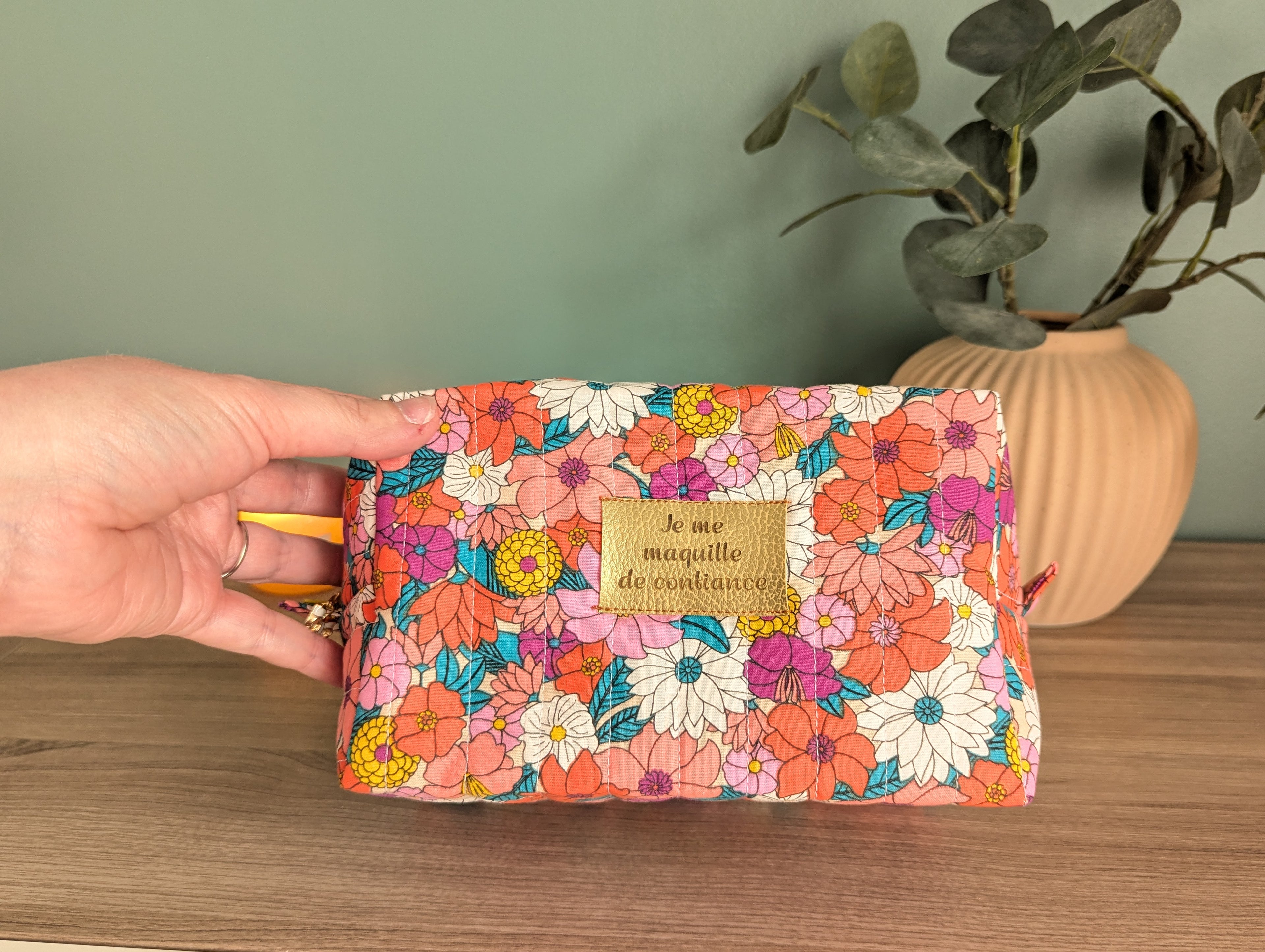 A hand holding a colorful floral makeup bag with a gold-colored tag that says "Je me mouquette de contenu".