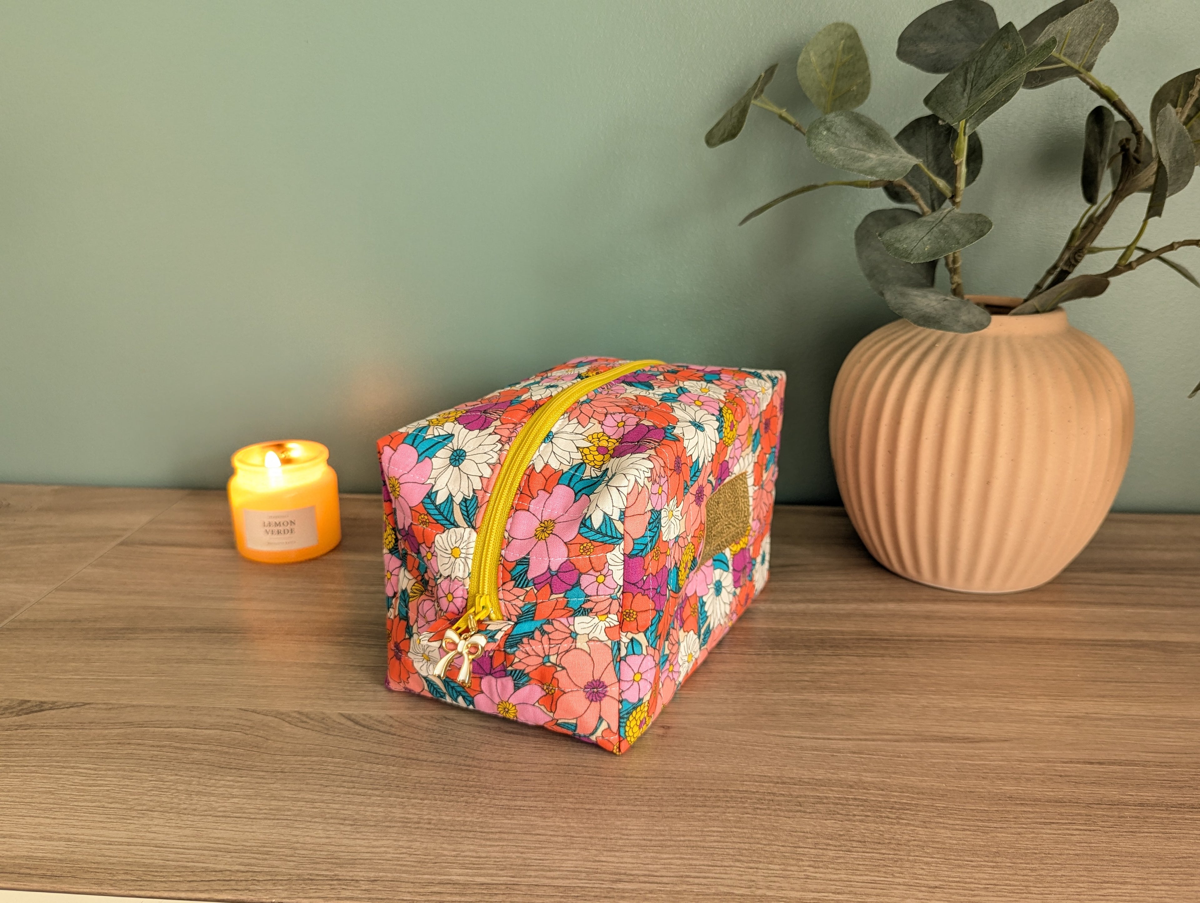 A floral-patterned makeup bag sits on a wooden surface next to a small candle and a vase with greenery.