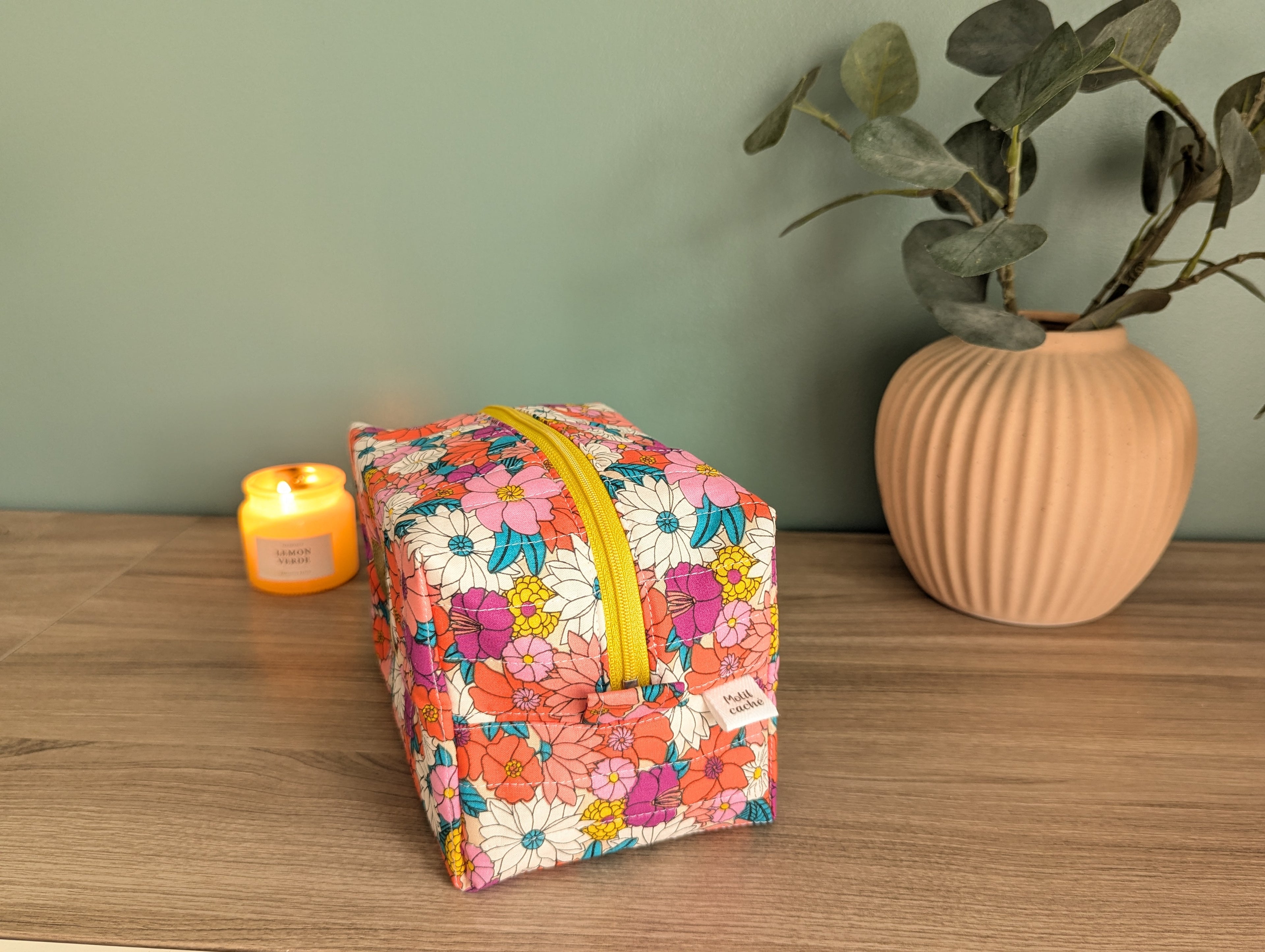 A floral-patterned makeup bag with a yellow zipper sits on a wooden surface, accompanied by a small candle and a potted plant.