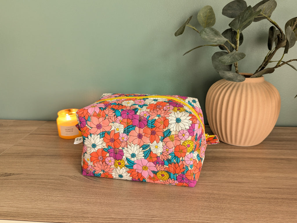 A colorful floral bag sits on a wooden surface, accompanied by a small yellow candle and a potted plant.