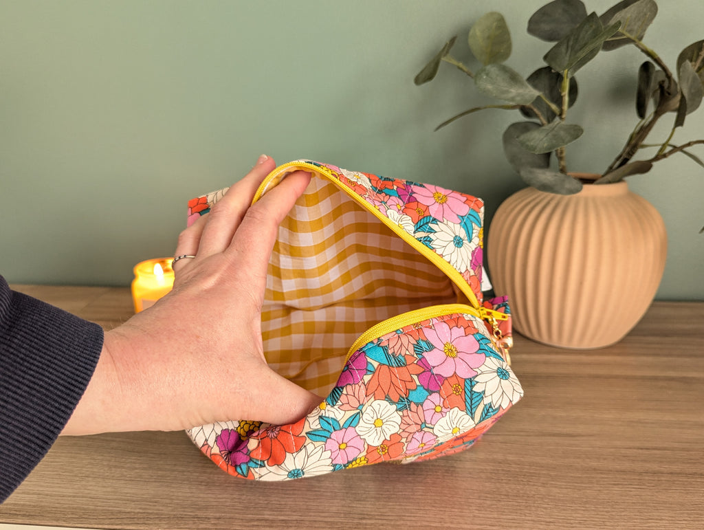 A hand is holding an open floral-patterned makeup bag on a wooden surface, with a potted plant and a lit candle in the background.