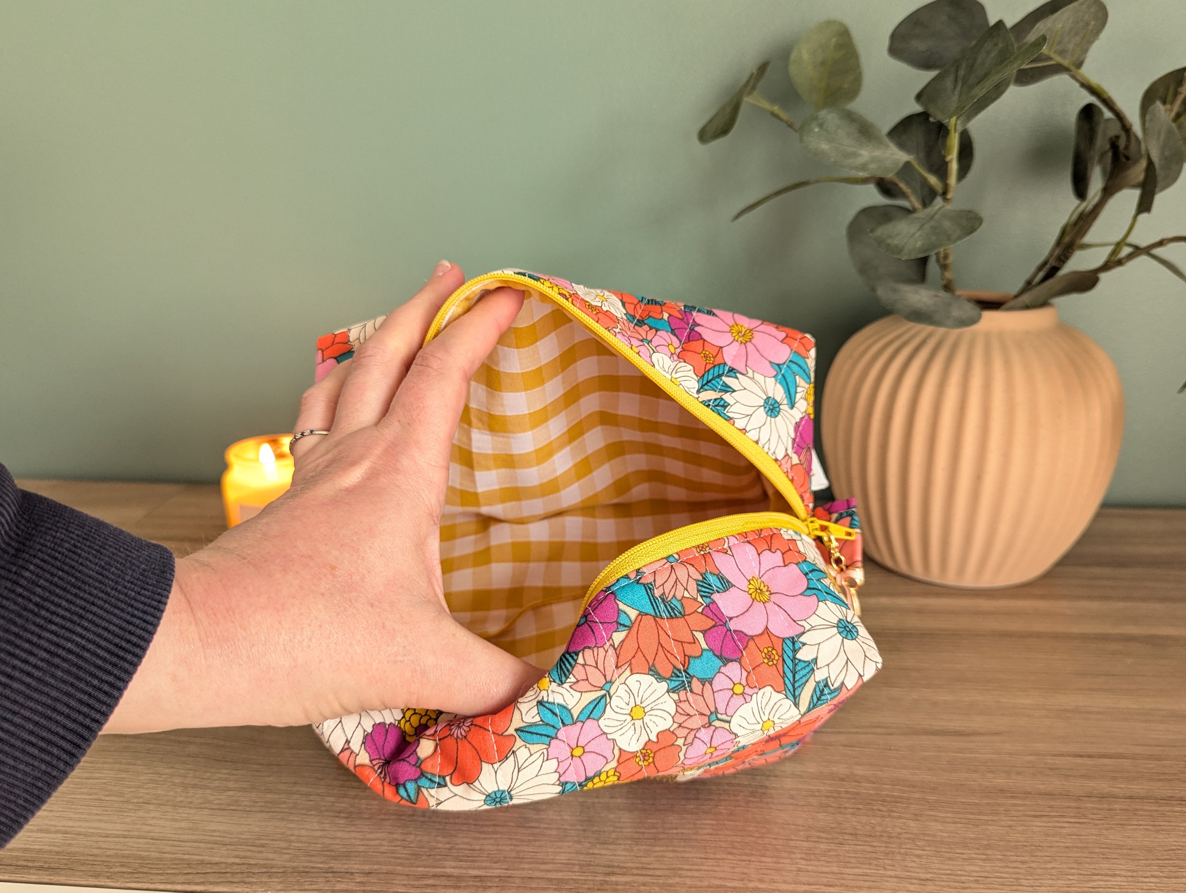 A hand is holding an open floral-patterned makeup bag on a wooden surface, with a potted plant and a lit candle in the background.