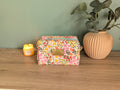 A floral-patterned makeup bag sits on a wooden surface next to a candle and a potted plant, creating a cozy and inviting atmosphere.