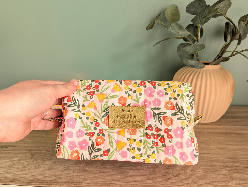 A hand holding a colorful floral makeup bag with a gold tag that says "le me MAJESTIE de l'examen".