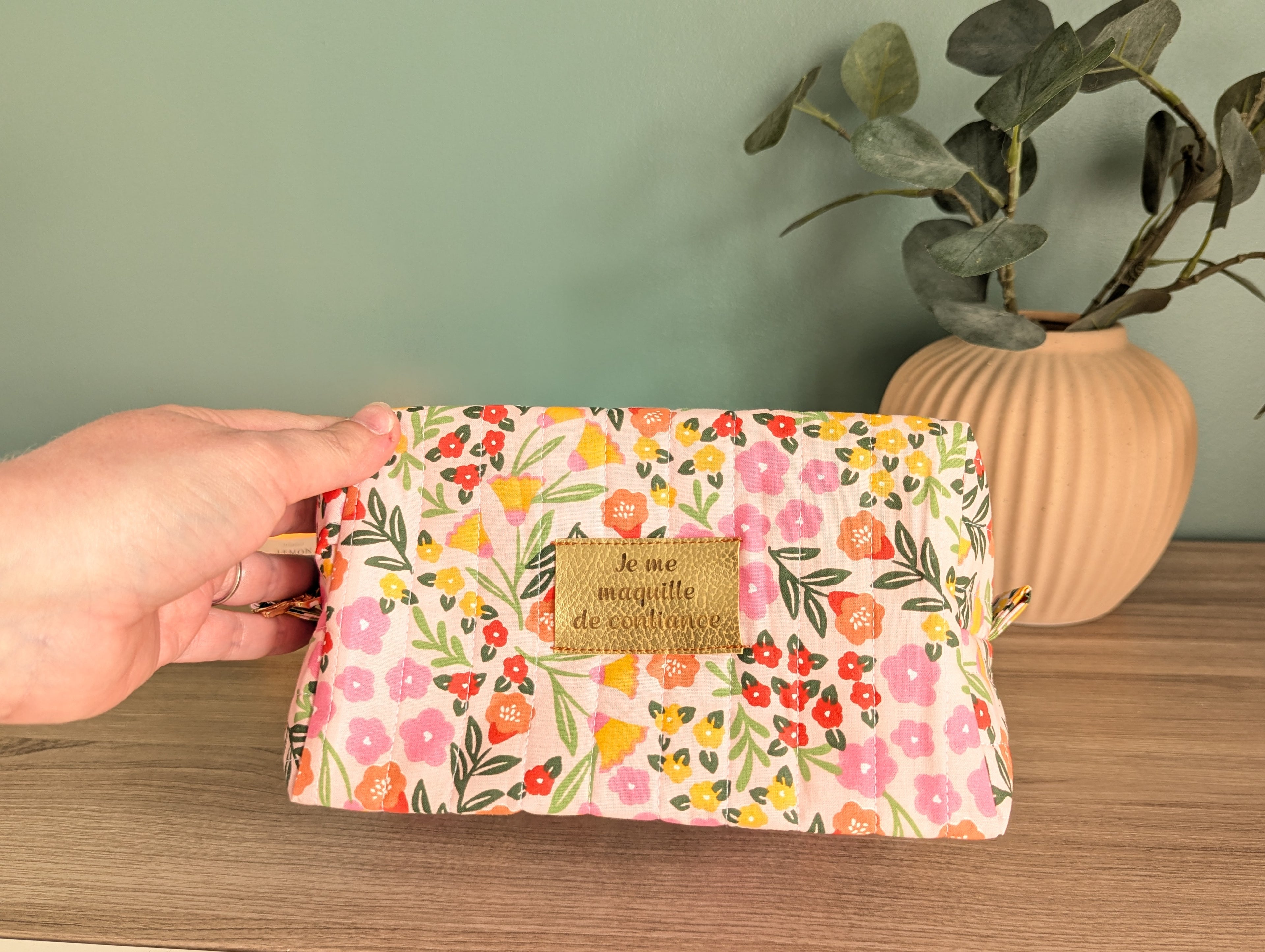 A hand holding a colorful floral makeup bag with a gold tag that says "le me MAJESTIE de l'examen".