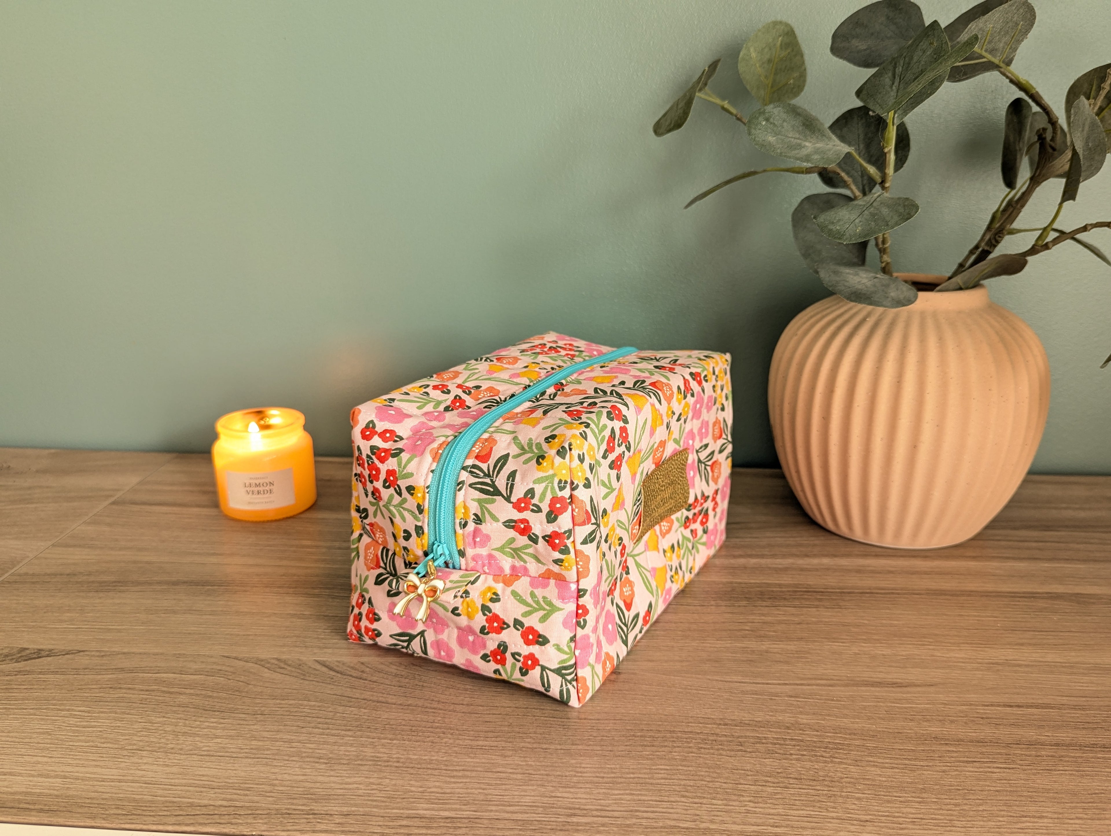 A floral-patterned makeup bag with a zipper sits on a wooden surface, accompanied by a small yellow candle and a potted plant.