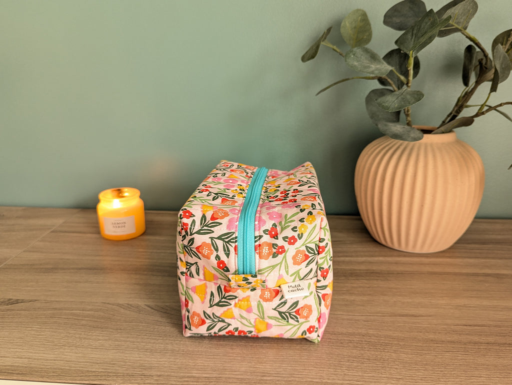 A floral-patterned makeup bag with a zipper sits on a wooden surface, accompanied by a small yellow candle and a potted plant.