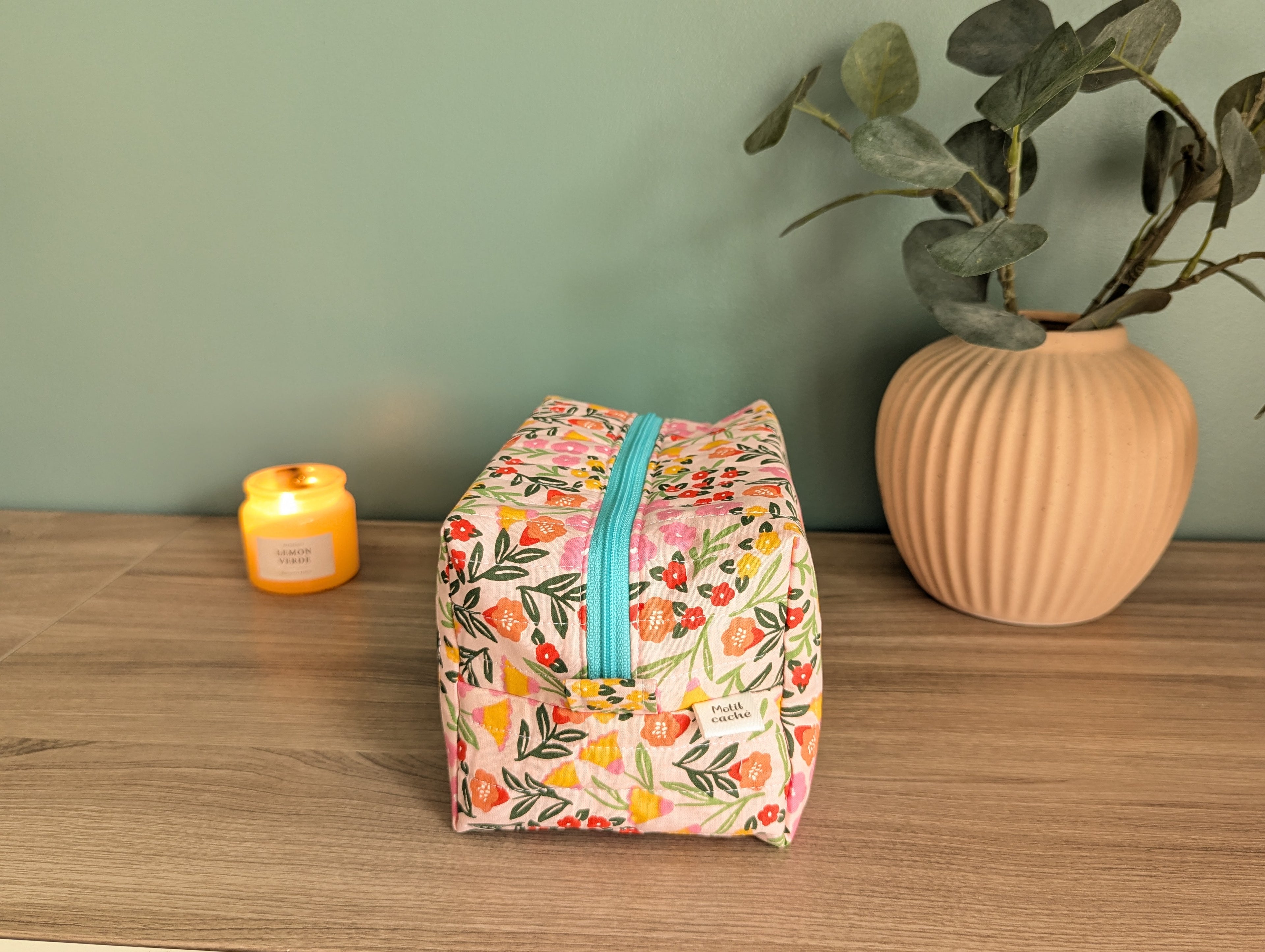 A floral-patterned makeup bag with a zipper sits on a wooden surface, accompanied by a small yellow candle and a potted plant.