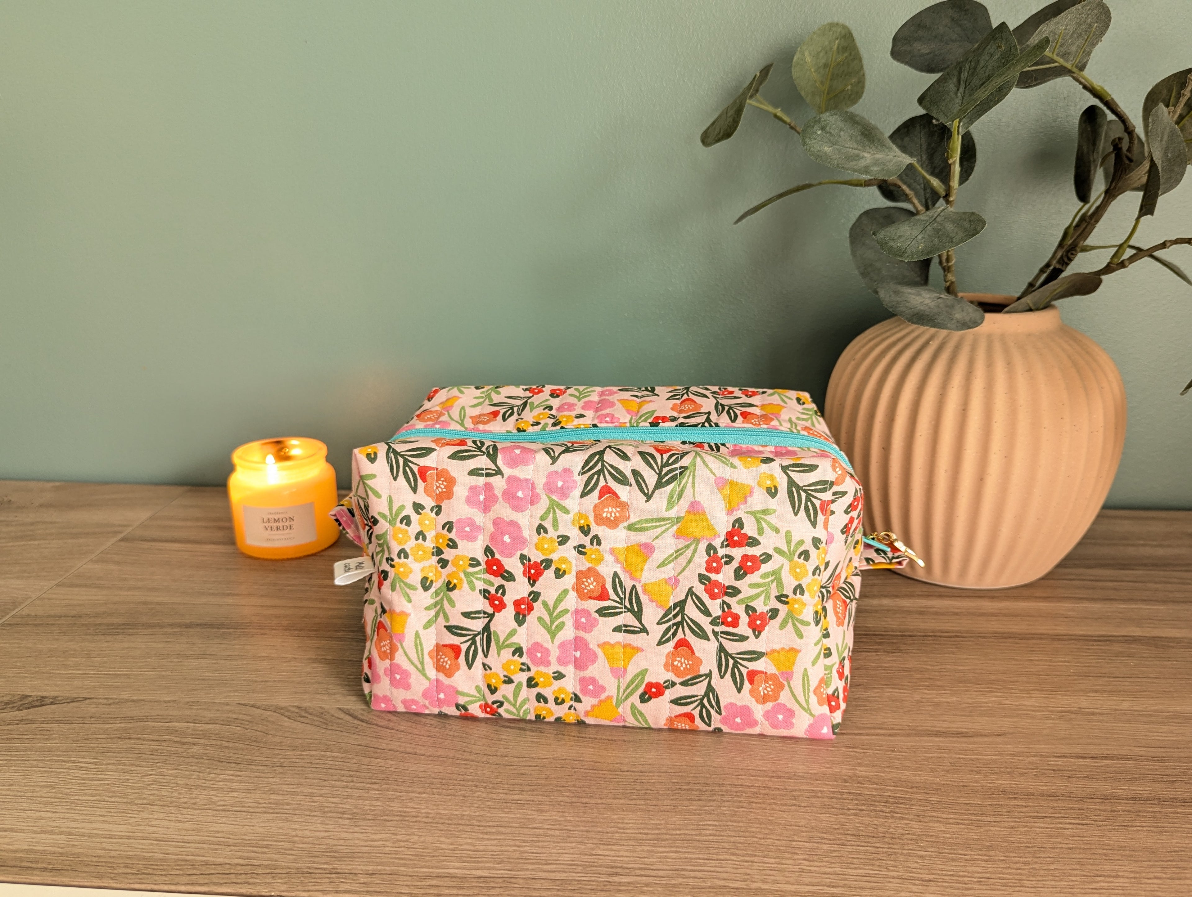 A floral patterned bag sits on a wooden surface, accompanied by a candle and a potted plant.
