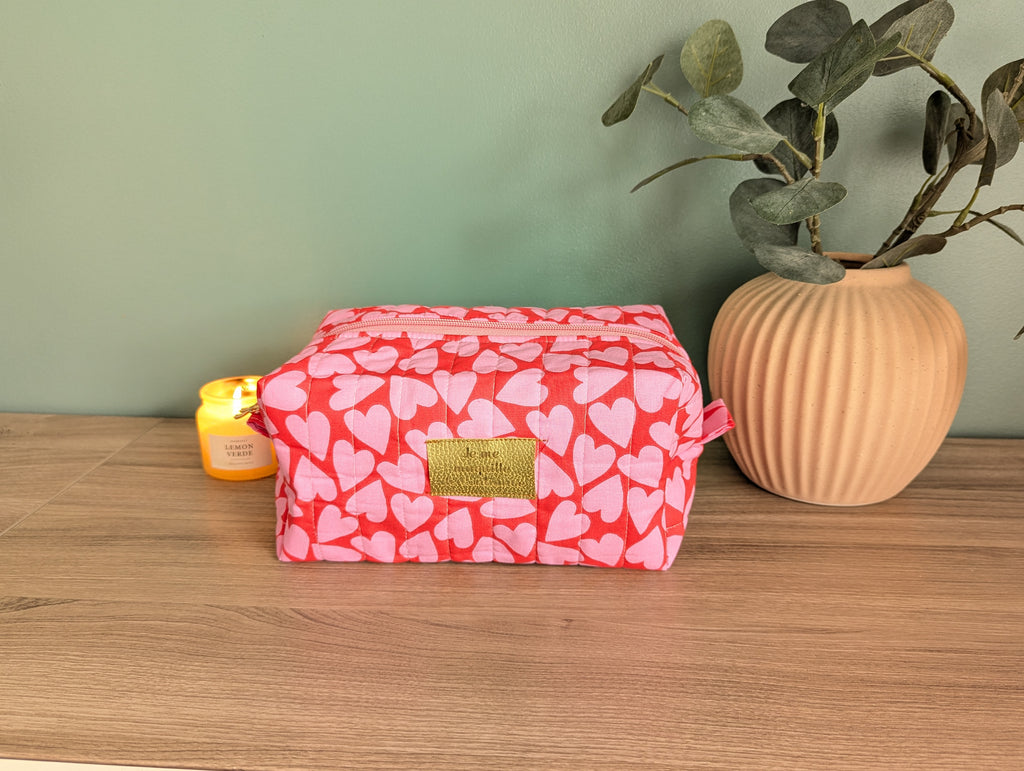 A red and pink heart-patterned cosmetic bag sits on a wooden surface, accompanied by a small yellow candle and a potted plant.