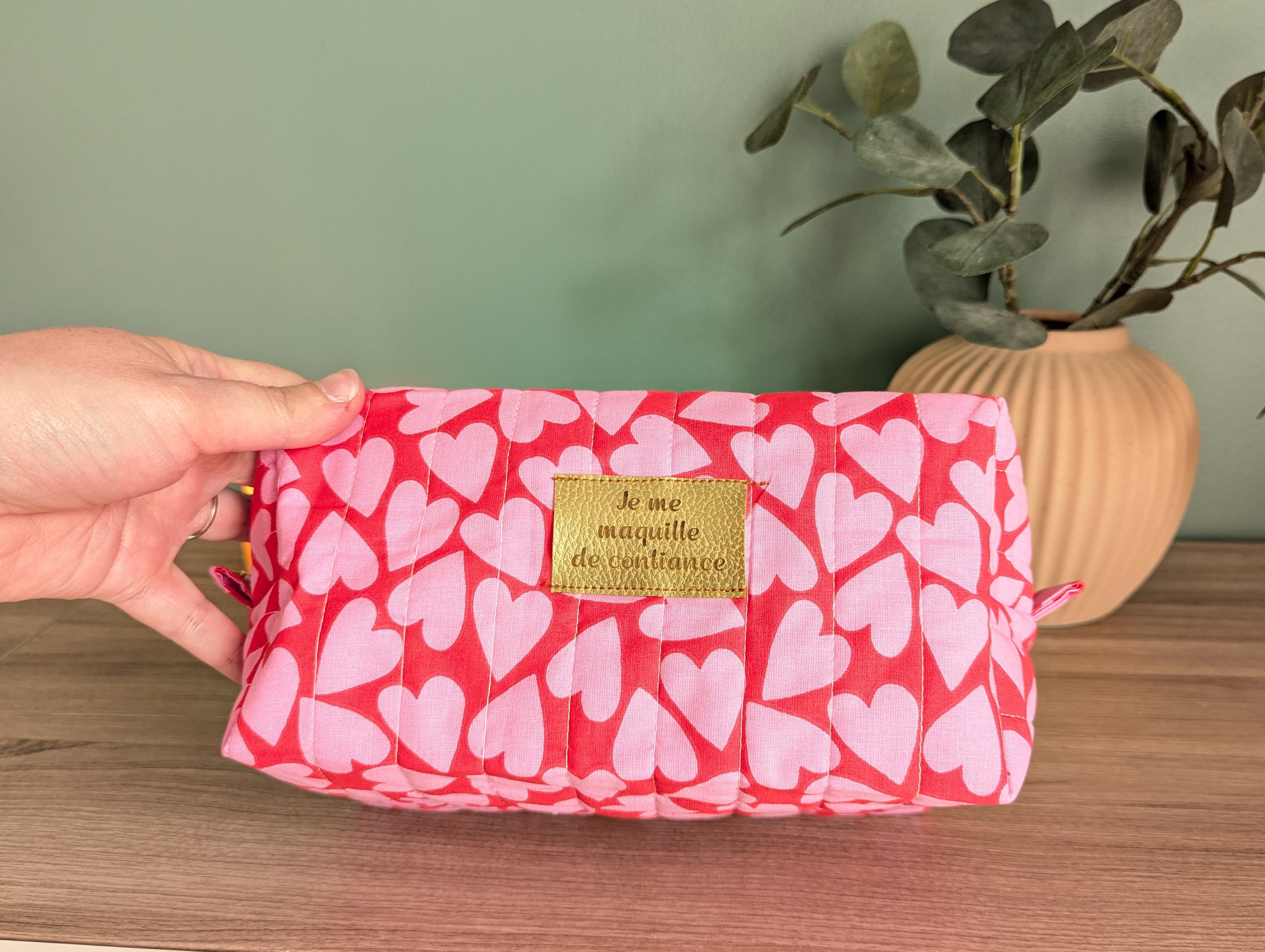 A hand is holding a pink makeup bag with a heart pattern, placed on a wooden surface next to a potted plant.