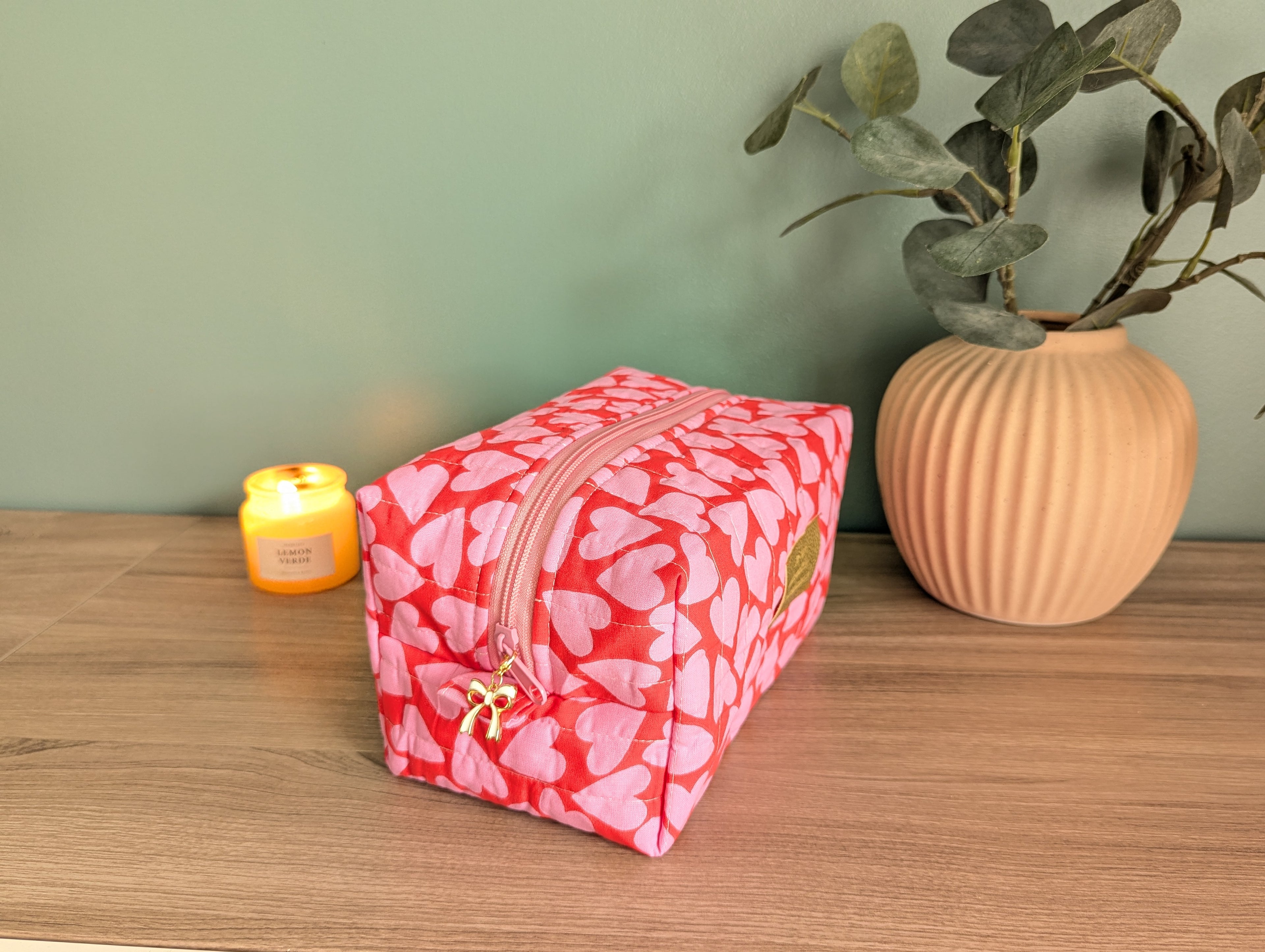 A pink and red heart-patterned cosmetic bag sits on a wooden surface, accompanied by a small yellow candle and a potted plant.