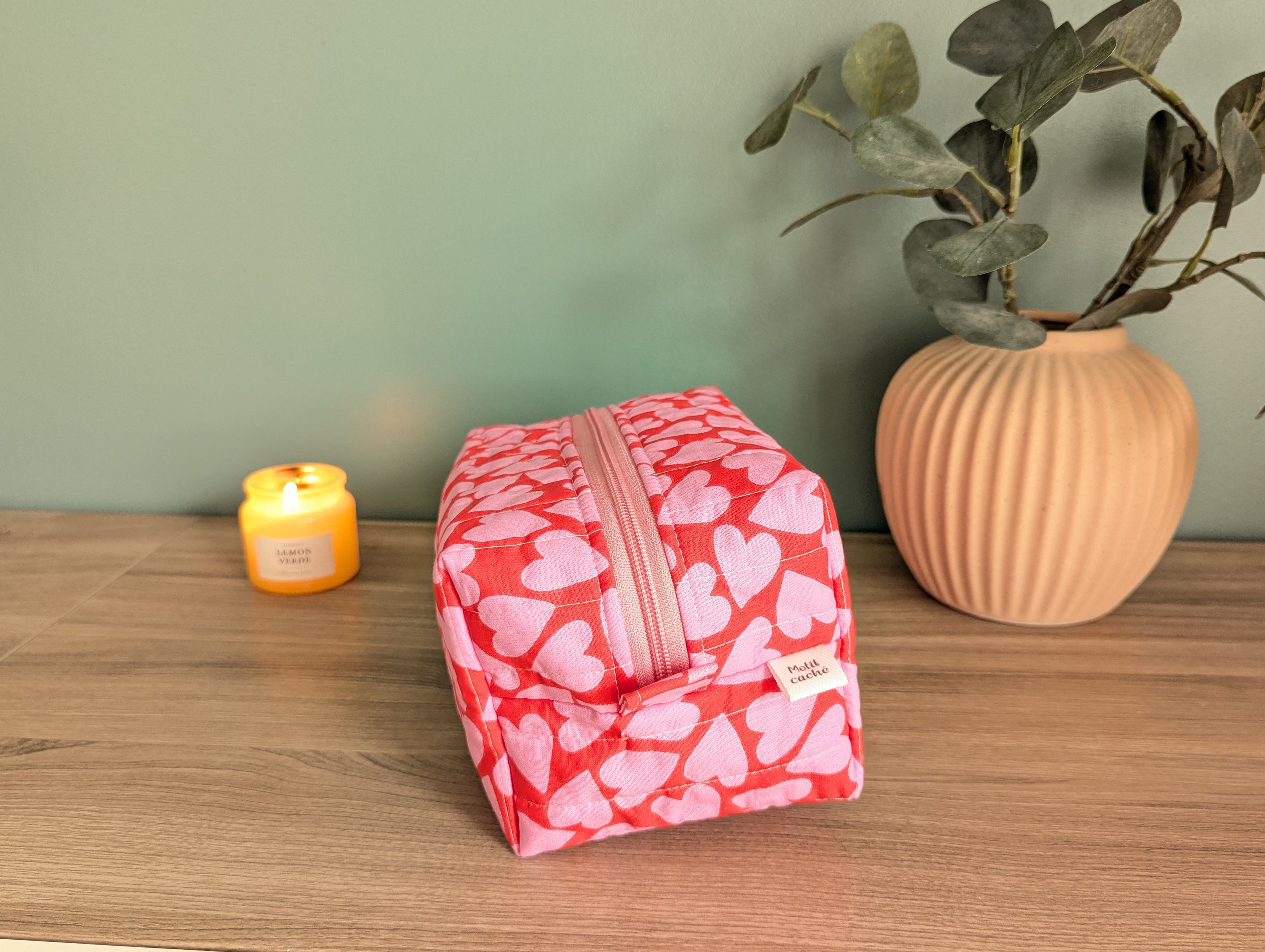 A red and white heart-patterned makeup bag sits on a wooden surface, accompanied by a small yellow candle and a potted plant.