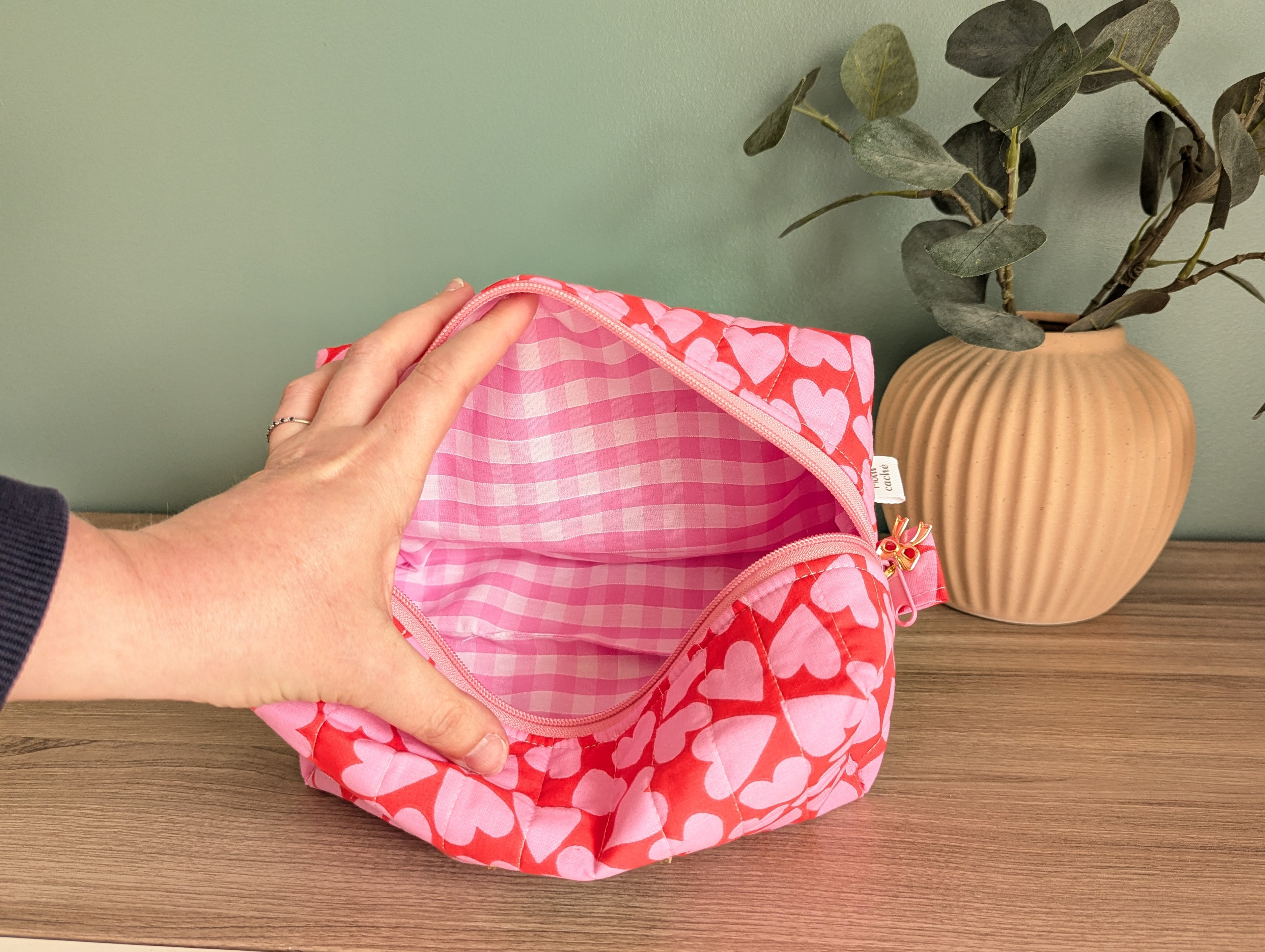 A hand is holding an open pink and white checkered bag with a heart pattern, placed on a wooden surface next to a potted plant.