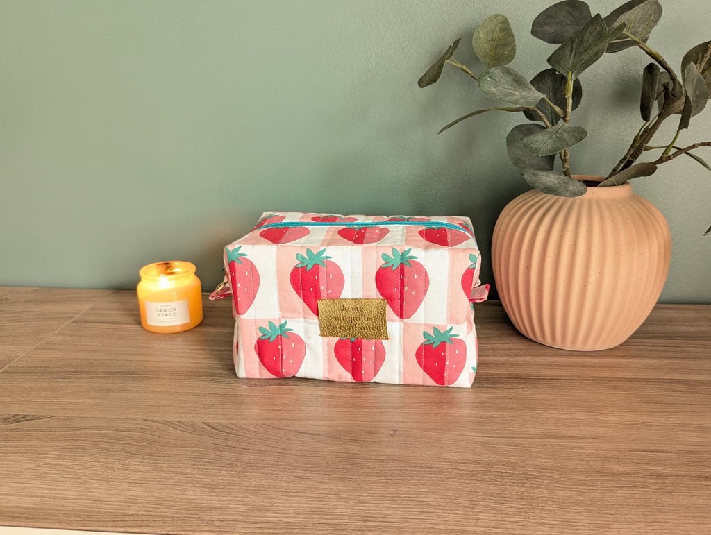 A wooden table with a pink vase, a candle, and a red and white striped bag with a strawberry pattern.