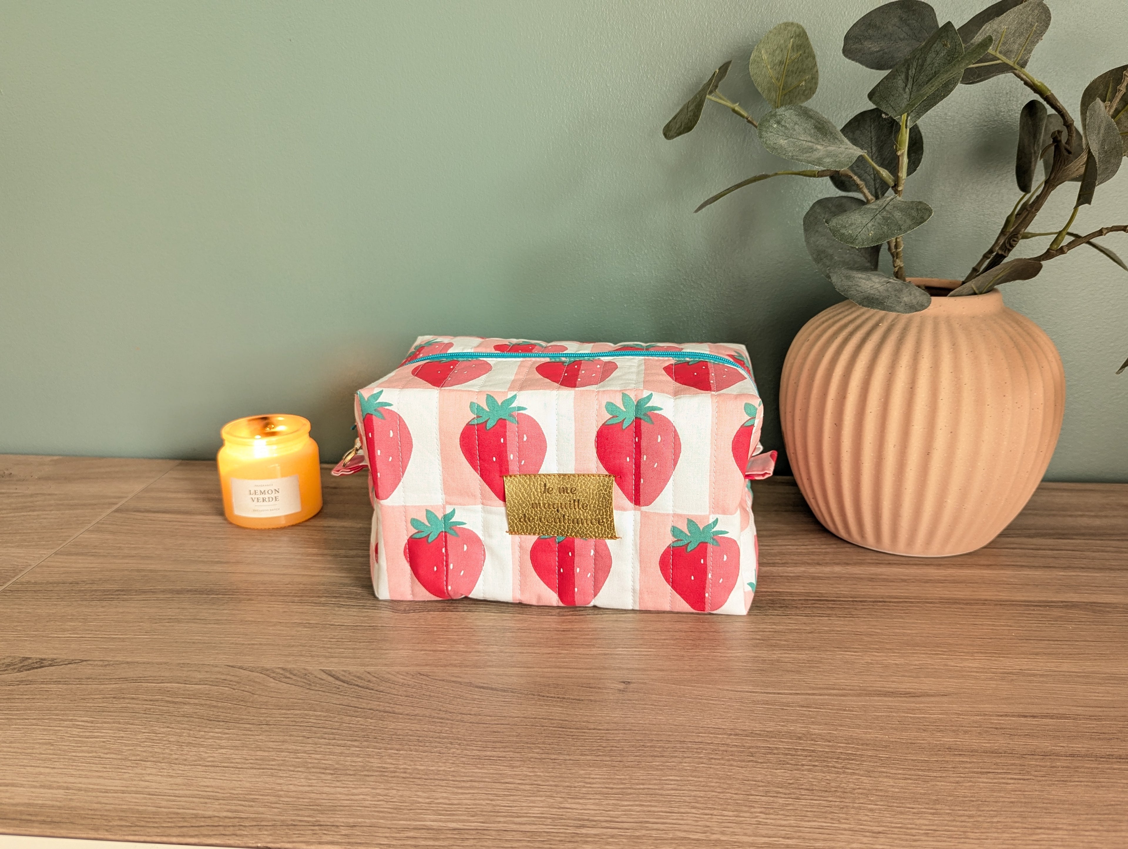 A wooden table with a pink vase, a candle, and a red and white striped bag with a strawberry pattern.