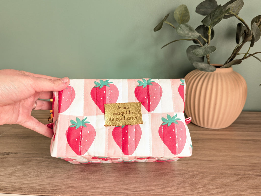 A hand is holding a small, colorful bag with a strawberry pattern, placed on a wooden surface next to a potted plant.