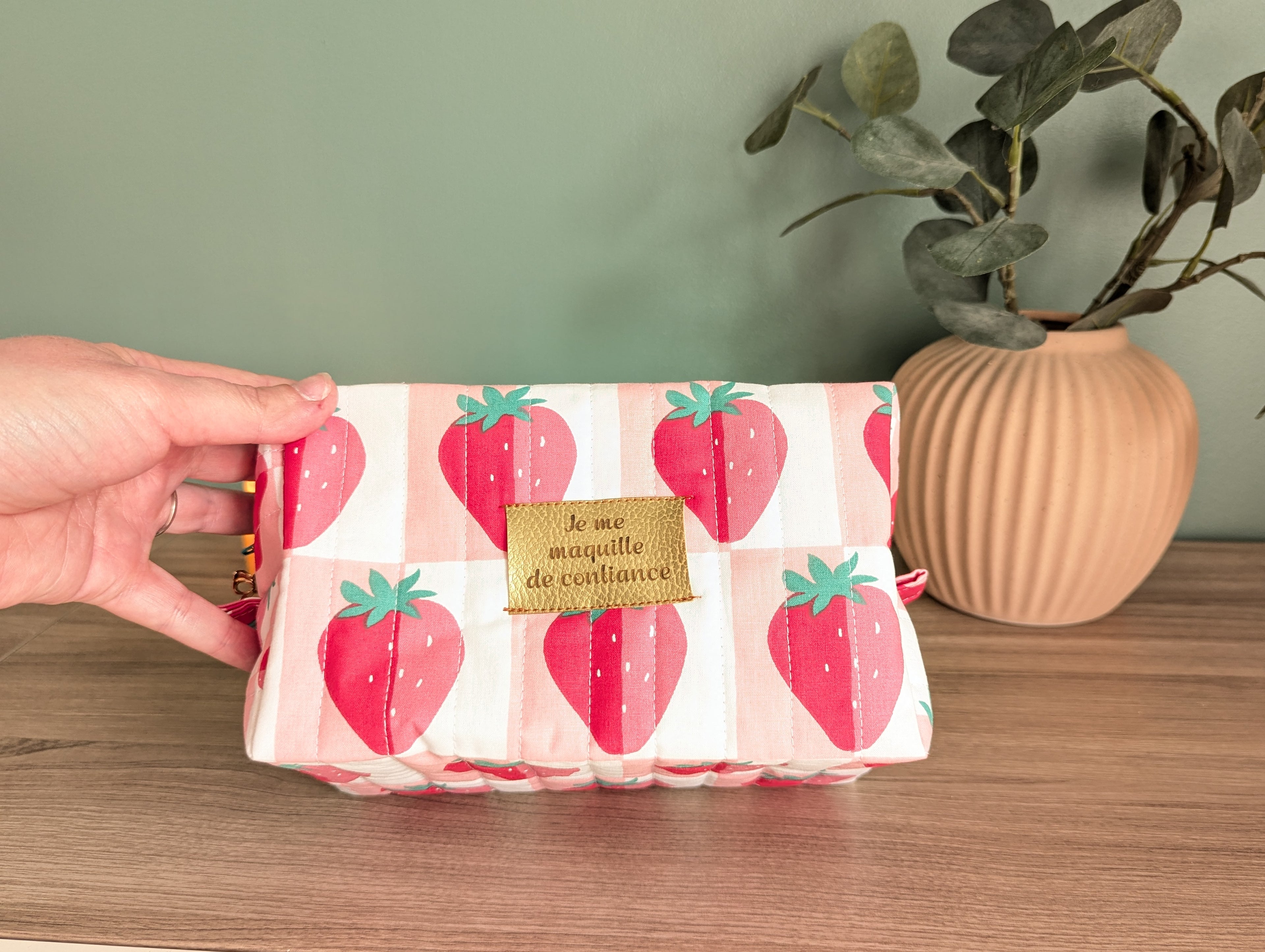 A hand is holding a small, colorful bag with a strawberry pattern, placed on a wooden surface next to a potted plant.