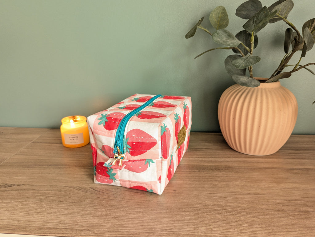 A red and white striped makeup bag with a blue zipper sits on a wooden surface, accompanied by a small yellow candle and a potted plant.