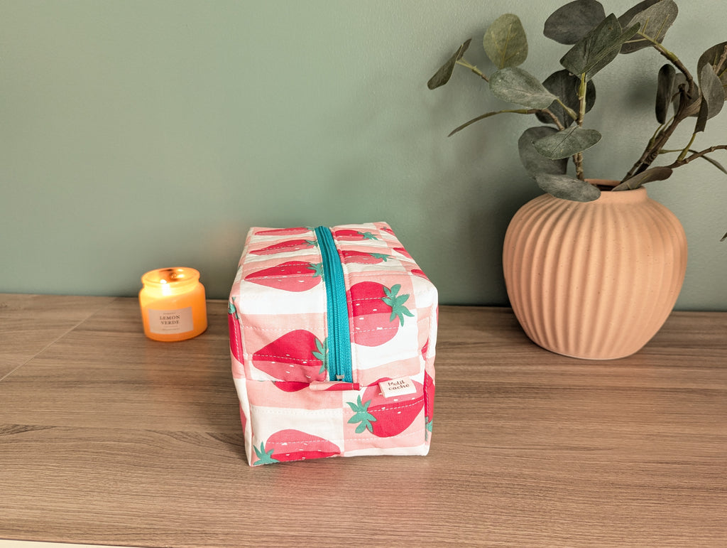 A pink and white striped cosmetic bag with a teal zipper is placed on a wooden surface next to a small orange candle and a potted plant.