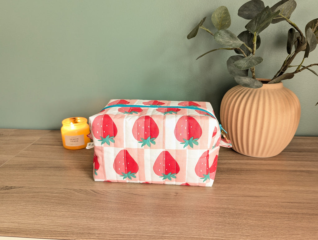 A wooden table with a white and red striped bag featuring a strawberry pattern, a yellow candle, and a potted plant.
