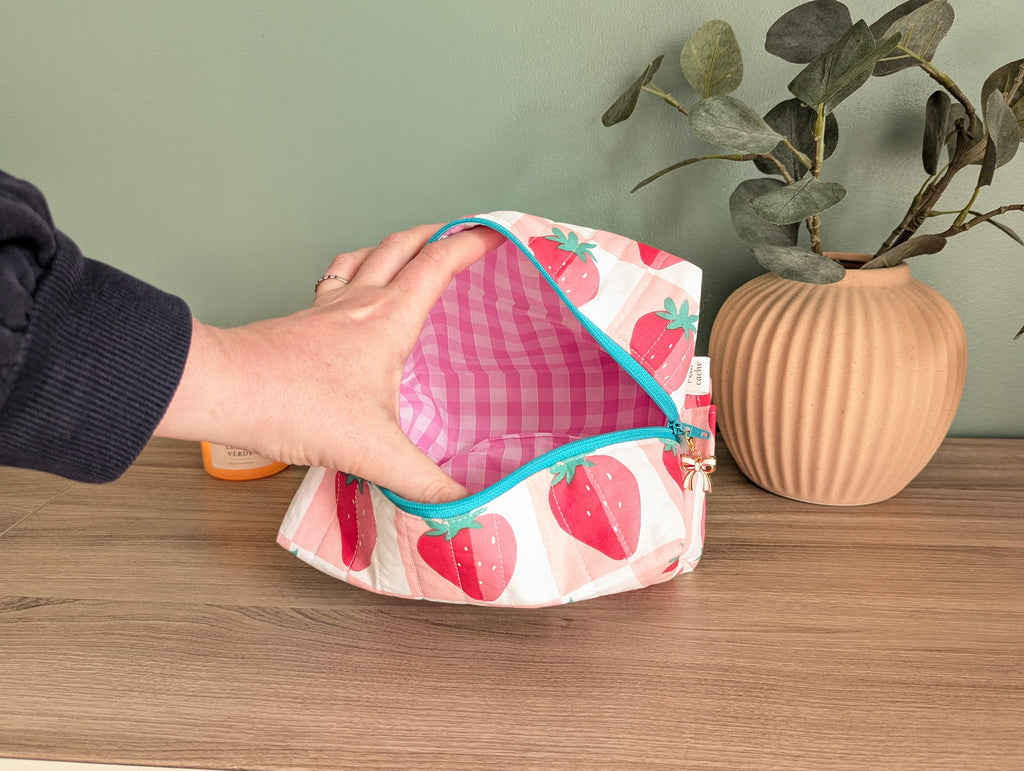 A hand is holding open a pink and white striped bag with a strawberry pattern, placed on a wooden surface next to a potted plant.
